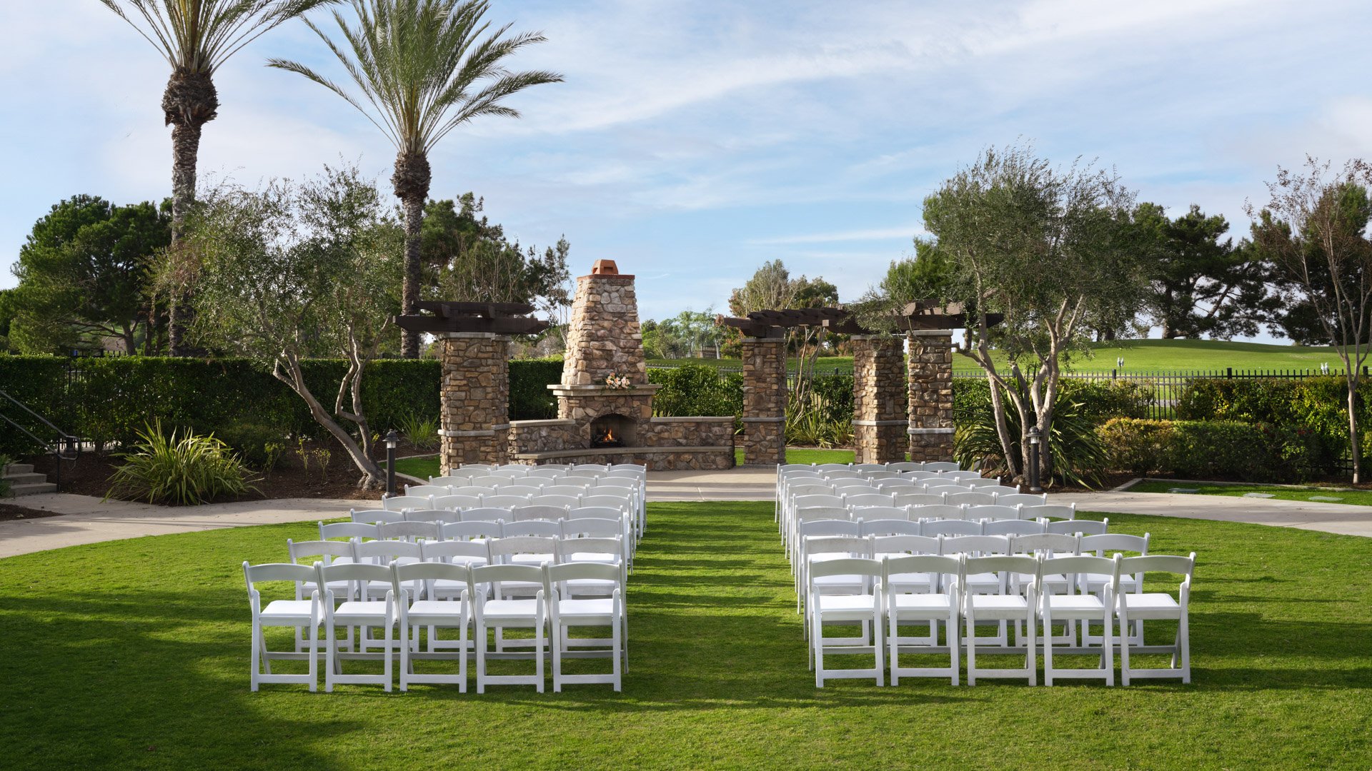 Timeless outdoor wedding ceremony with white folding chairs arranged on green lawn facing stone fireplace and pergola at Aliso Viejo by Wedgewood Weddings