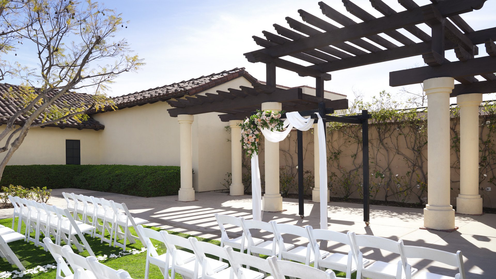 Intimate outdoor wedding ceremony with pergola, white fabric draping, floral arch, and white chairs at Aliso Viejo by Wedgewood Weddings venue