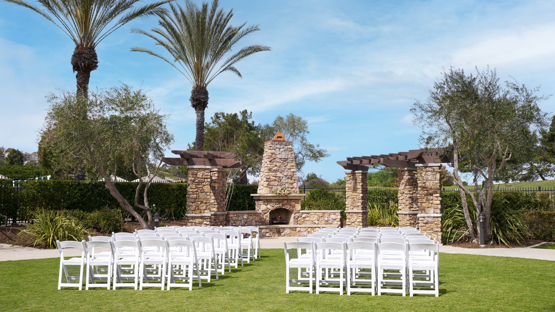 Picturesque outdoor wedding ceremony with white chairs, palm trees, stone fireplace backdrop, and blue sky at Aliso Viejo by Wedgewood Weddings venue