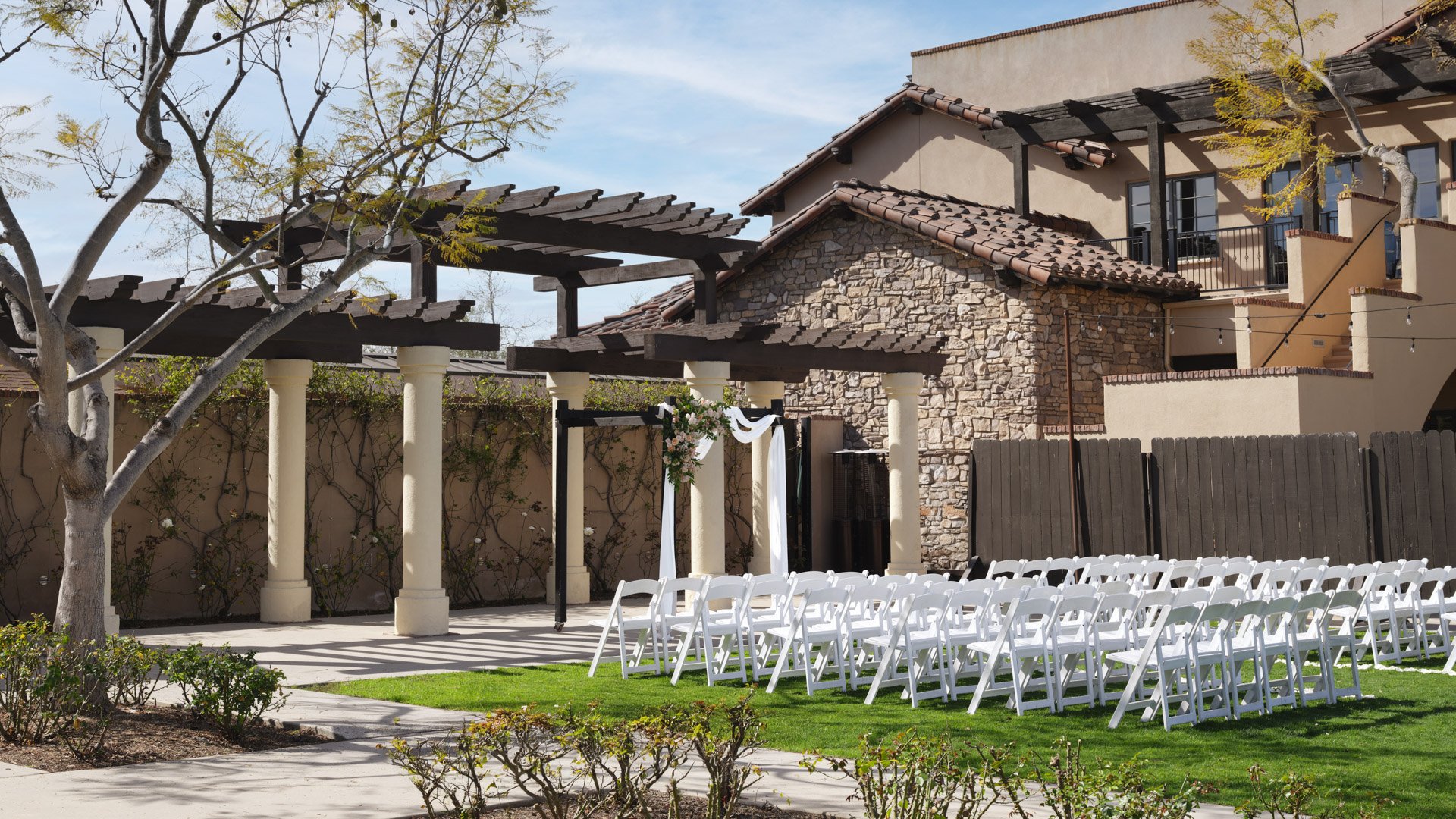 Scenic outdoor wedding ceremony featuring wooden pergola with floral arrangements, white chairs, and lush landscaping at Aliso Viejo by Wedgewood Weddings California
