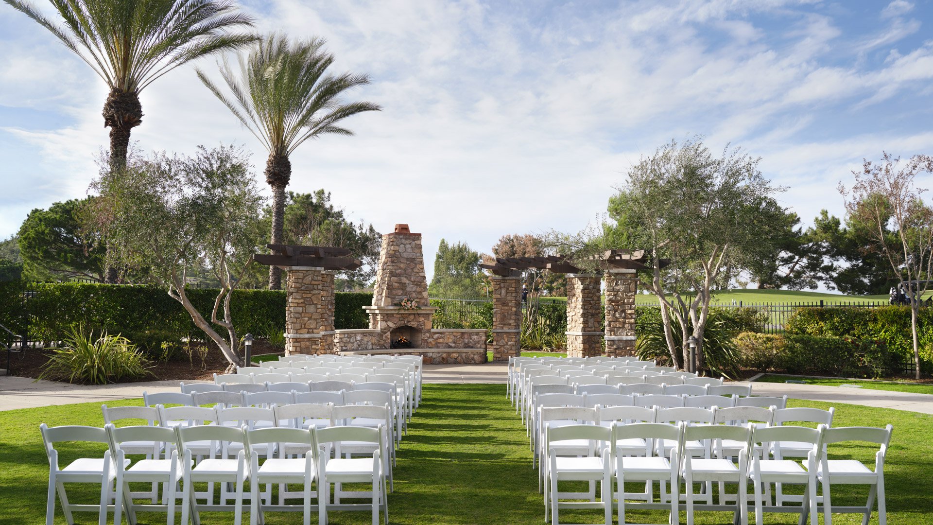 Beautiful garden wedding ceremony with white folding chairs, manicured lawn, and stone fireplace backdrop at Aliso Viejo by Wedgewood Weddings California