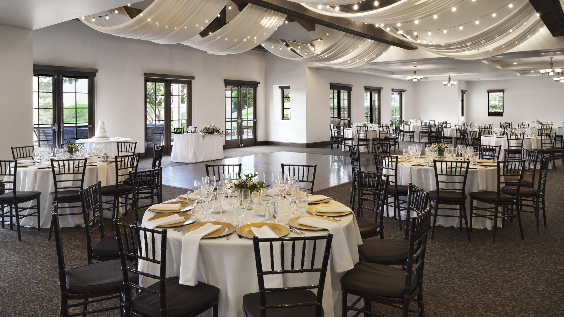 Spacious wedding reception ballroom featuring elegant ceiling draping with string lights, round tables with gold chargers, and black Chiavari chairs at Aliso Viejo by Wedgewood Weddings