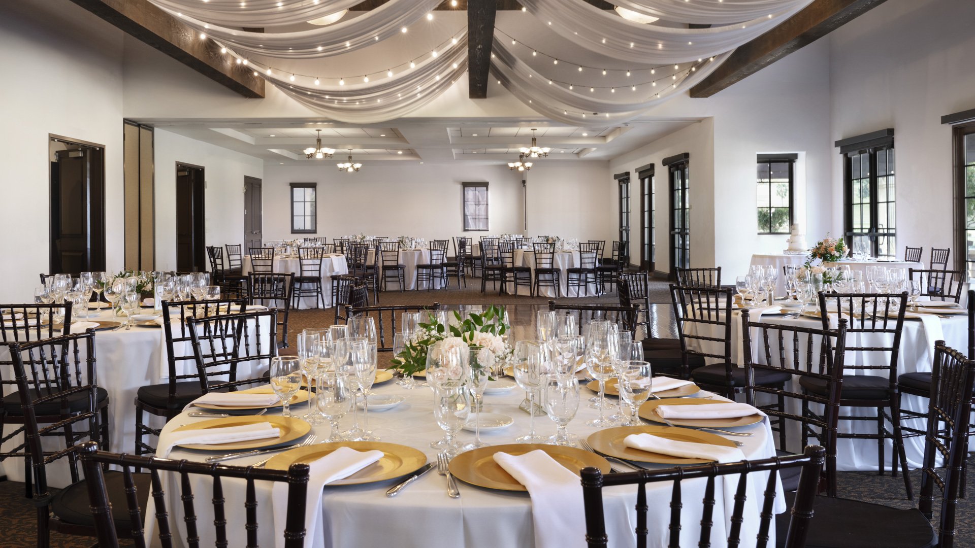 Elegant indoor wedding reception ballroom with round tables, gold chargers, black Chiavari chairs, and draped ceiling with string lights at Aliso Viejo venue