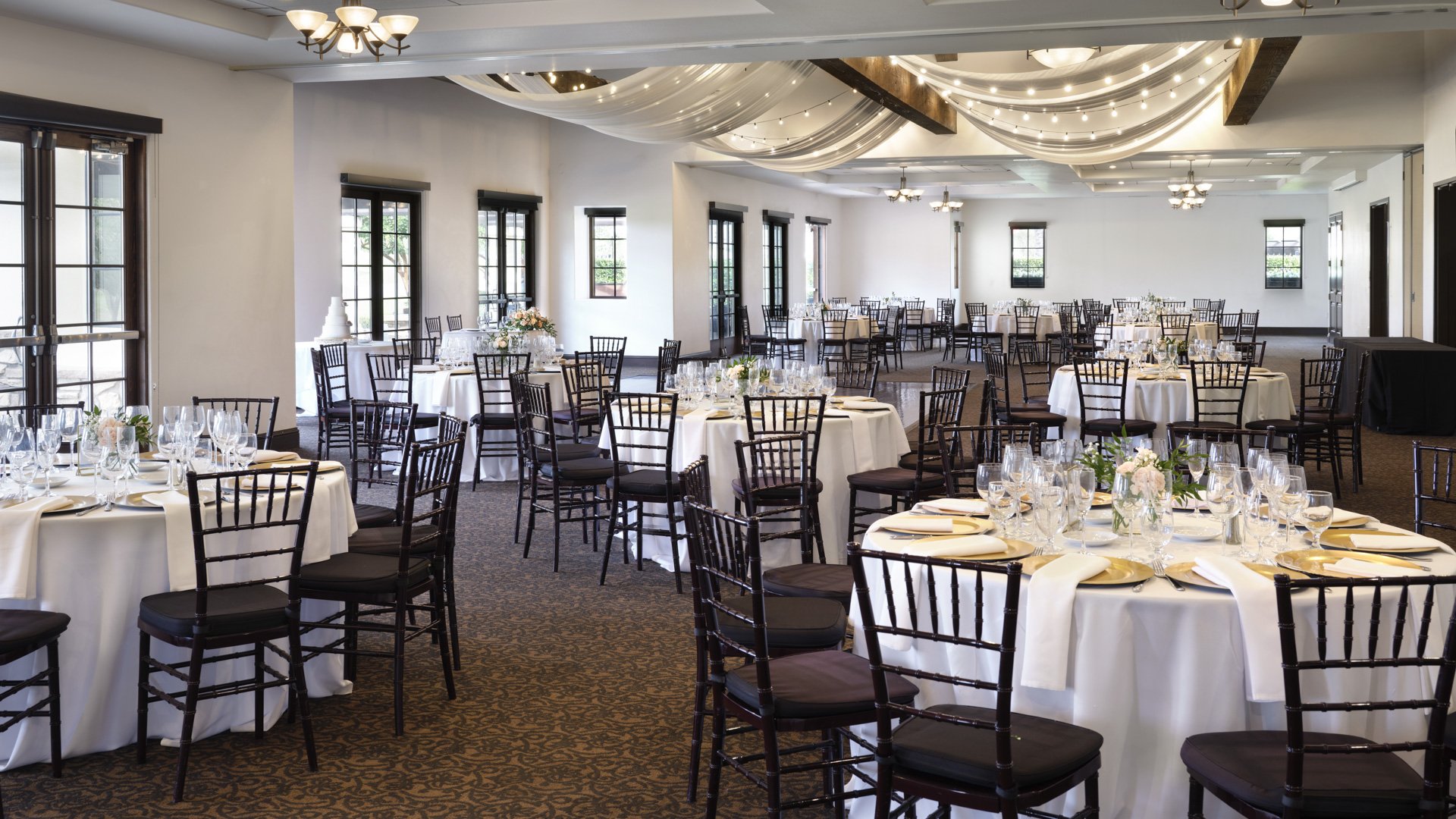 Elegant indoor wedding reception with draped ceiling, string lights, round tables with gold place settings, and black Chiavari chairs at Aliso Viejo California venue