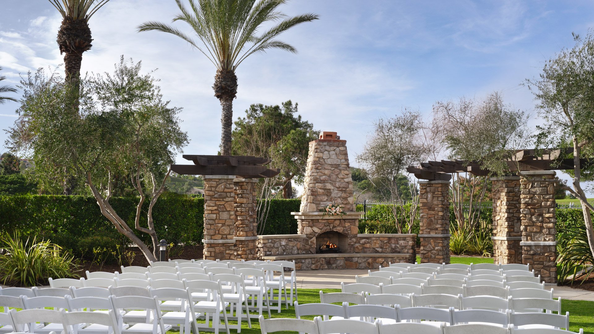 Elegant outdoor wedding ceremony with white chairs arranged on green lawn facing stone fireplace altar at Aliso Viejo by Wedgewood Weddings venue in California