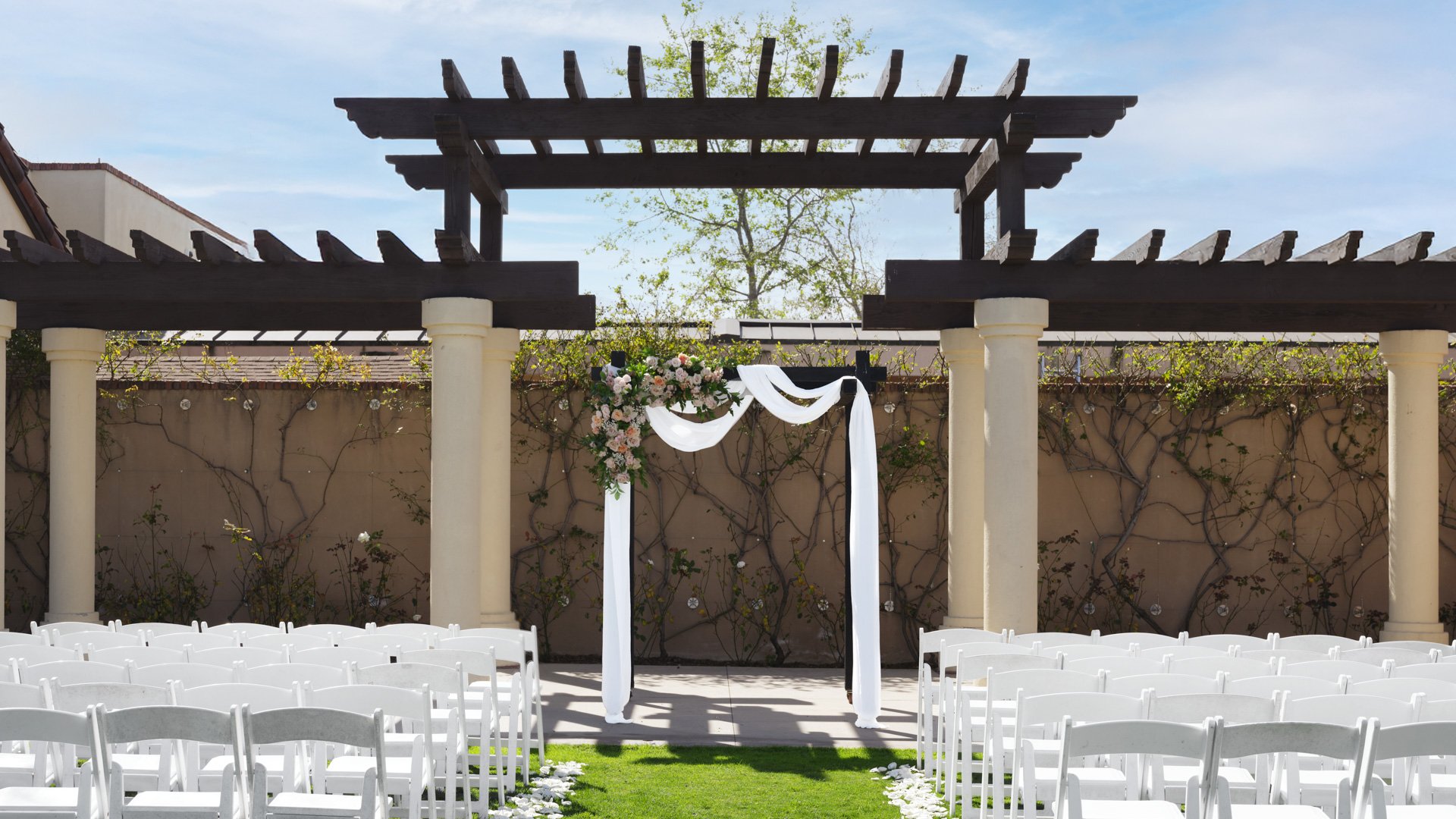Beautiful outdoor wedding ceremony under wooden pergola with white fabric draping, floral arch, and white folding chairs at Aliso Viejo by Wedgewood Weddings California