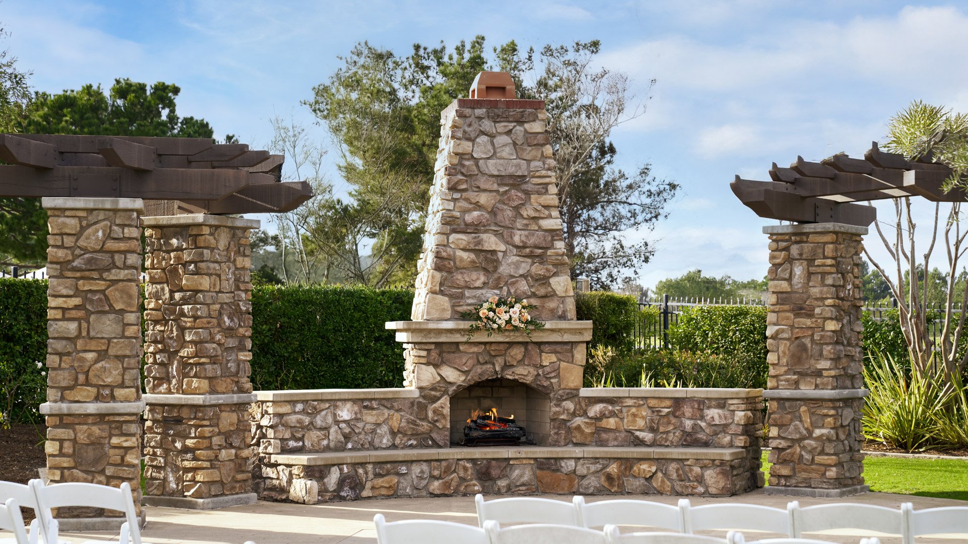 Stunning outdoor wedding ceremony with white chairs facing stone fireplace altar with wooden pergola structures at Aliso Viejo by Wedgewood Weddings California