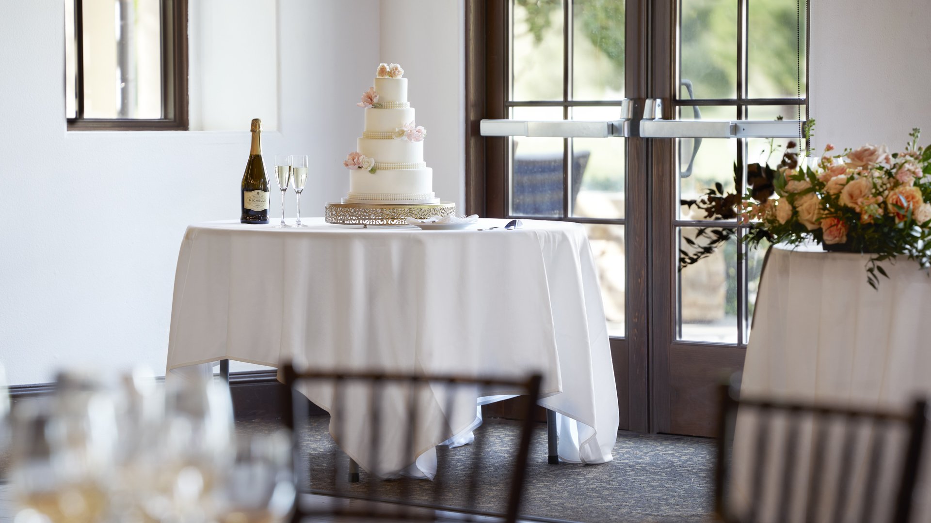Four-tier white wedding cake with pearl details on elegant table with champagne and floral arrangements at Aliso Viejo wedding venue