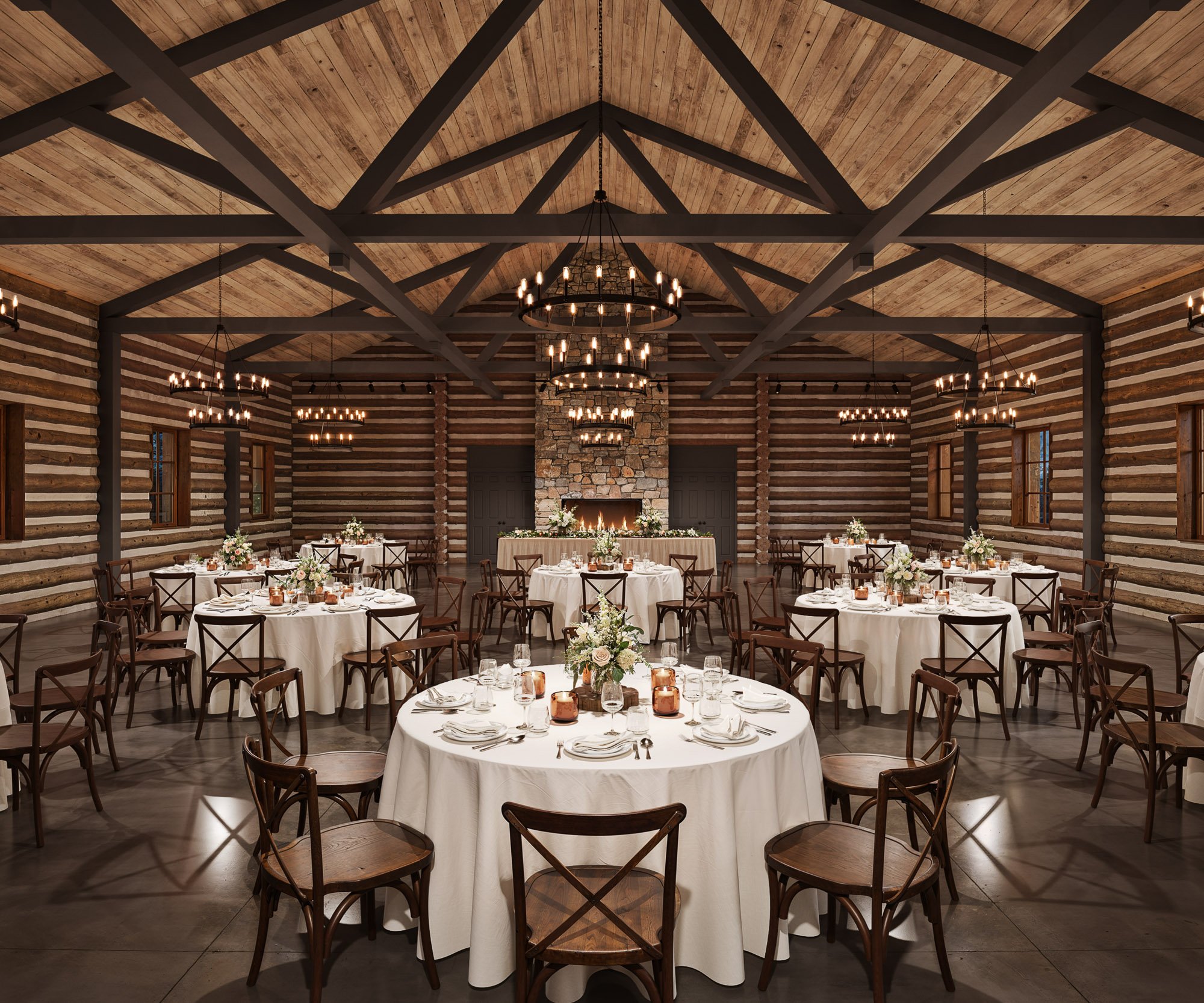 Chic log cabin wedding reception hall at Casa del Rio featuring wooden beam ceiling, stone fireplace, elegant chandeliers, and round tables with floral centerpieces.