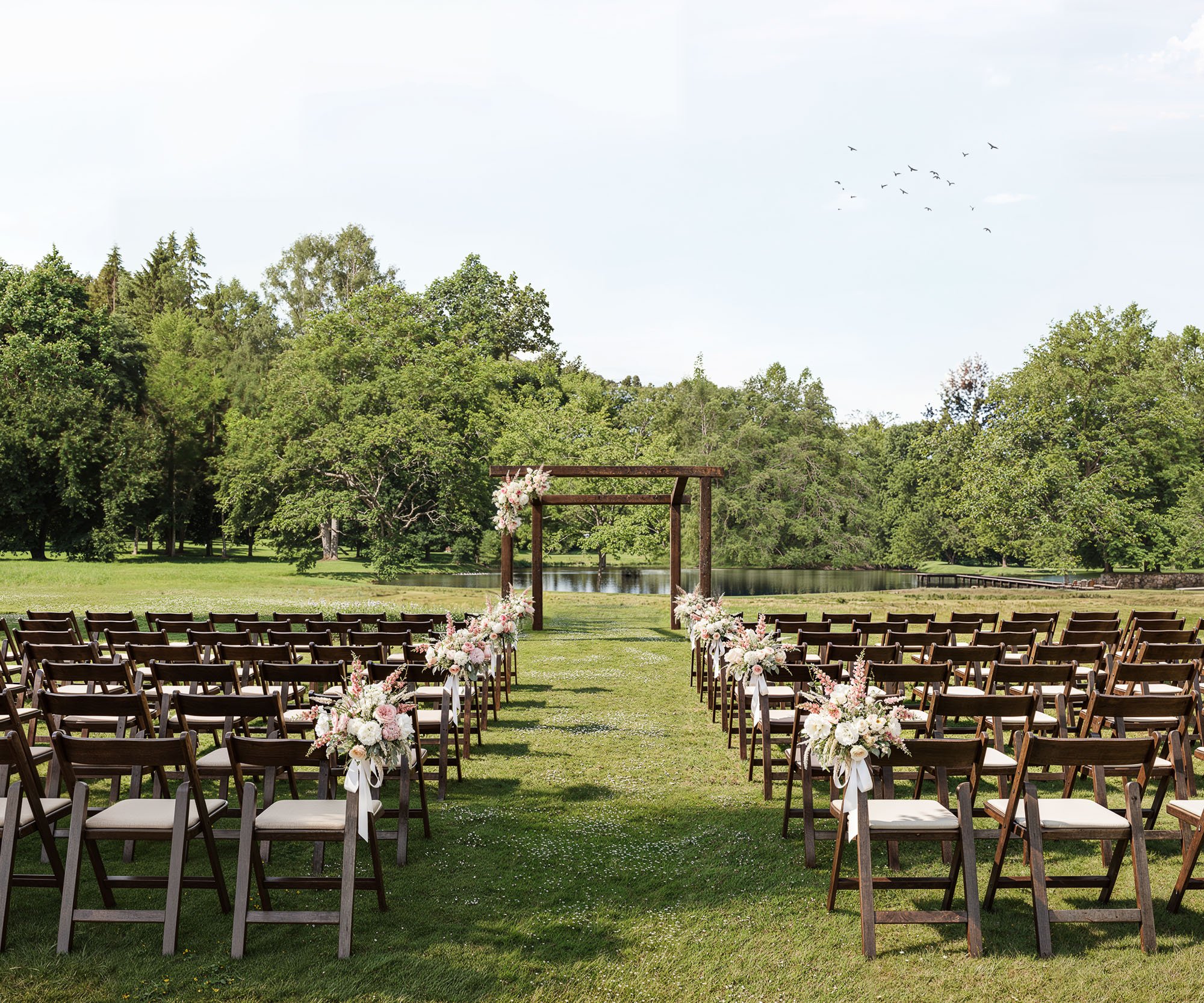 Serene lakeside wedding ceremony setup at Casa del Rio with wooden arch, floral decorations, and rows of chairs overlooking water surrounded by lush greenery.