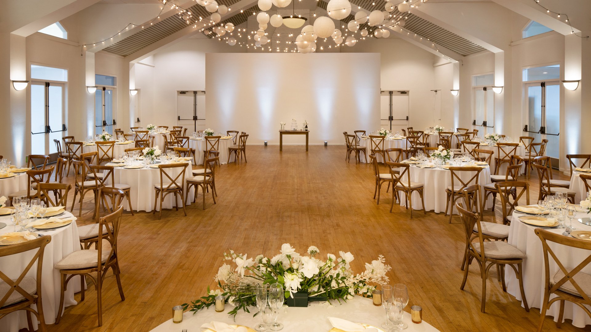 Spacious indoor wedding reception venue at Cuvier Club by Wedgewood Weddings featuring vaulted ceilings, string lights, paper lanterns, and cross-back chairs in La Jolla