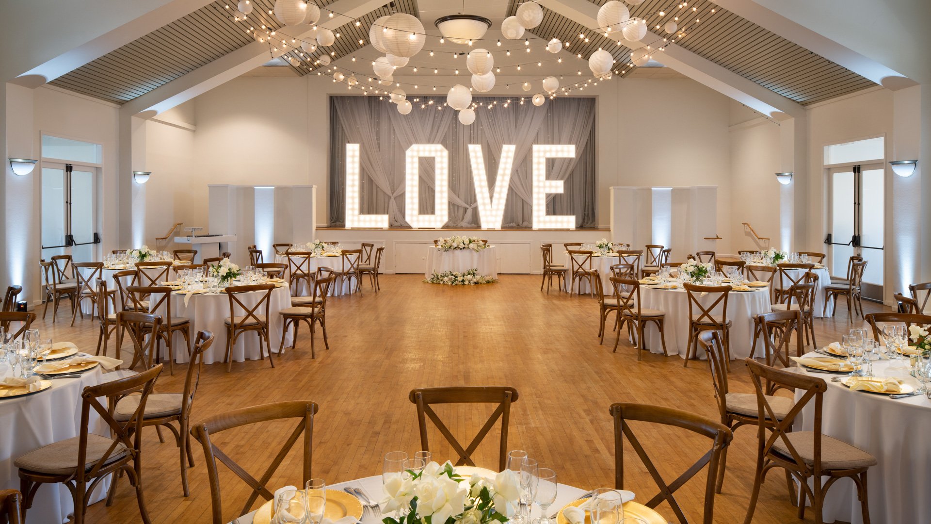 Sophisticated indoor wedding reception at Cuvier Club by Wedgewood Weddings featuring illuminated LOVE marquee sign, cross-back wooden chairs, white linens, paper lantern ceiling, and hardwood floors in La Jolla