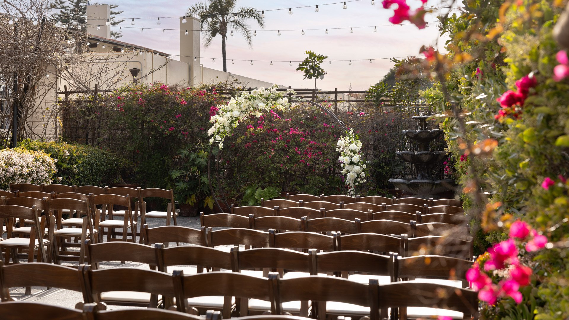 Beautiful Garden Courtyard wedding ceremony setup at Cuvier Club La Jolla with wooden chairs, white floral ceremony arch, colorful bougainvillea, and fountain backdrop
