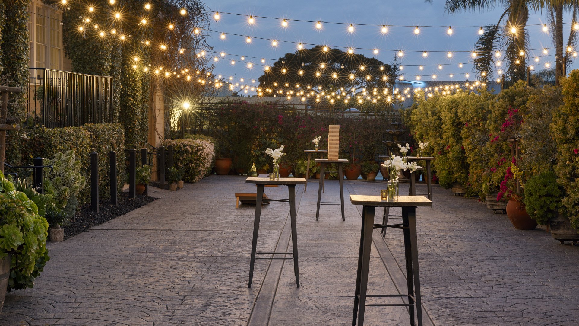 Romantic evening cocktail hour in the Garden Courtyard at Cuvier Club La Jolla with string bistro lights, high-top tables, white floral arrangements, and tropical landscaping