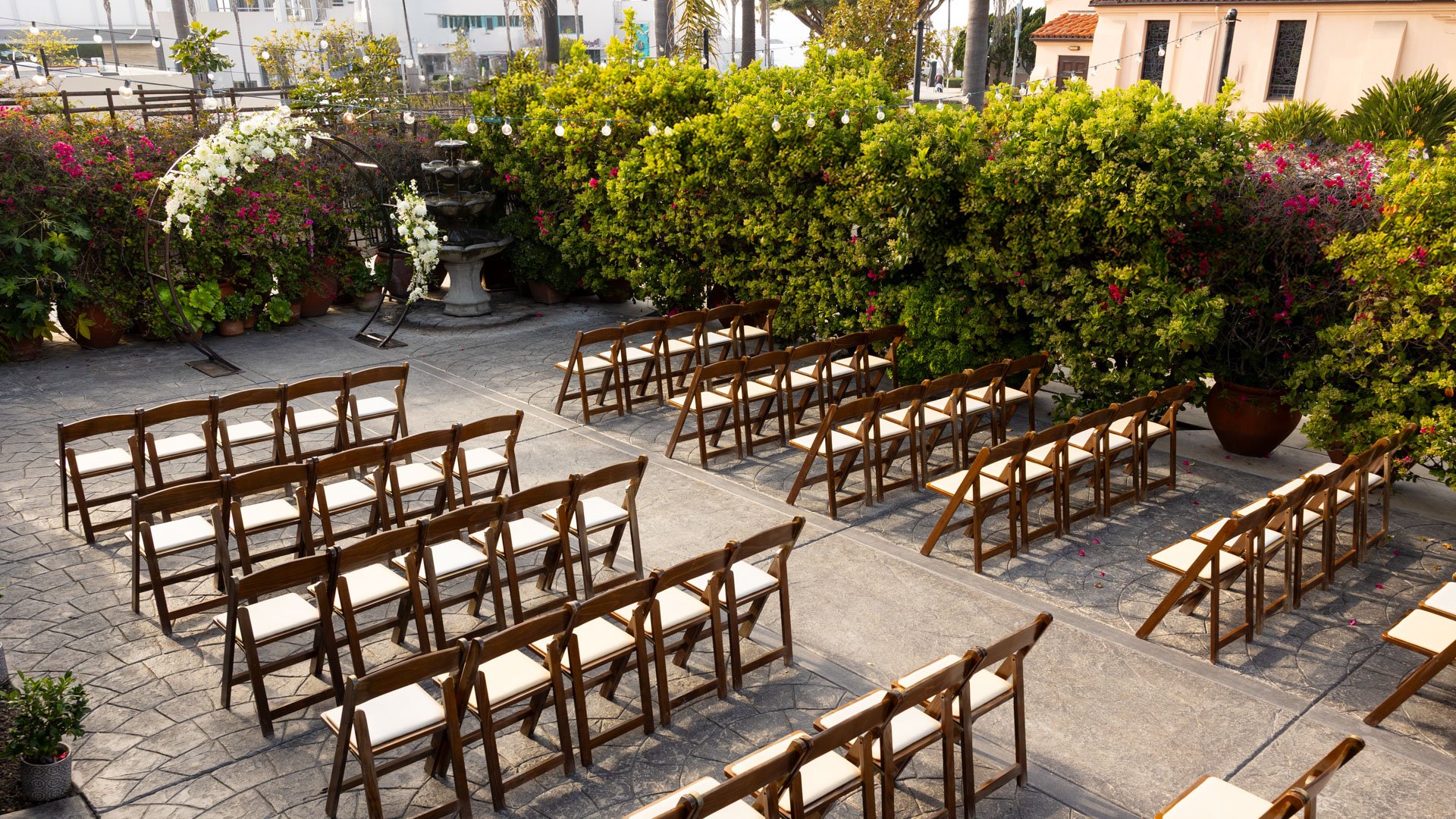 Intimate outdoor wedding ceremony space in the Garden Courtyard at Cuvier Club featuring wooden cross-back chairs, white floral arch, fountain backdrop, and vibrant bougainvillea blooms