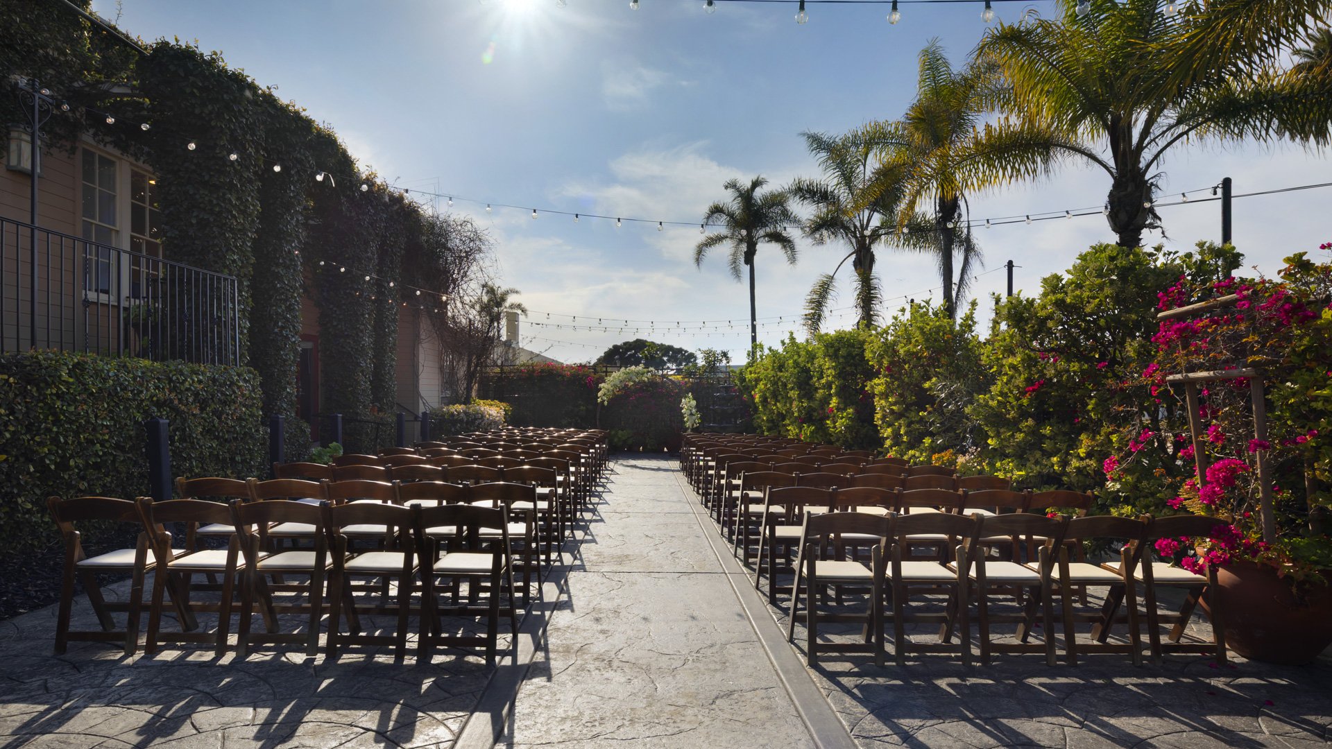 Beautiful Garden Courtyard ceremony space at Cuvier Club featuring wooden cross-back chairs arranged for guests, vibrant bougainvillea blooms, string lights, and tropical palm trees in La Jolla