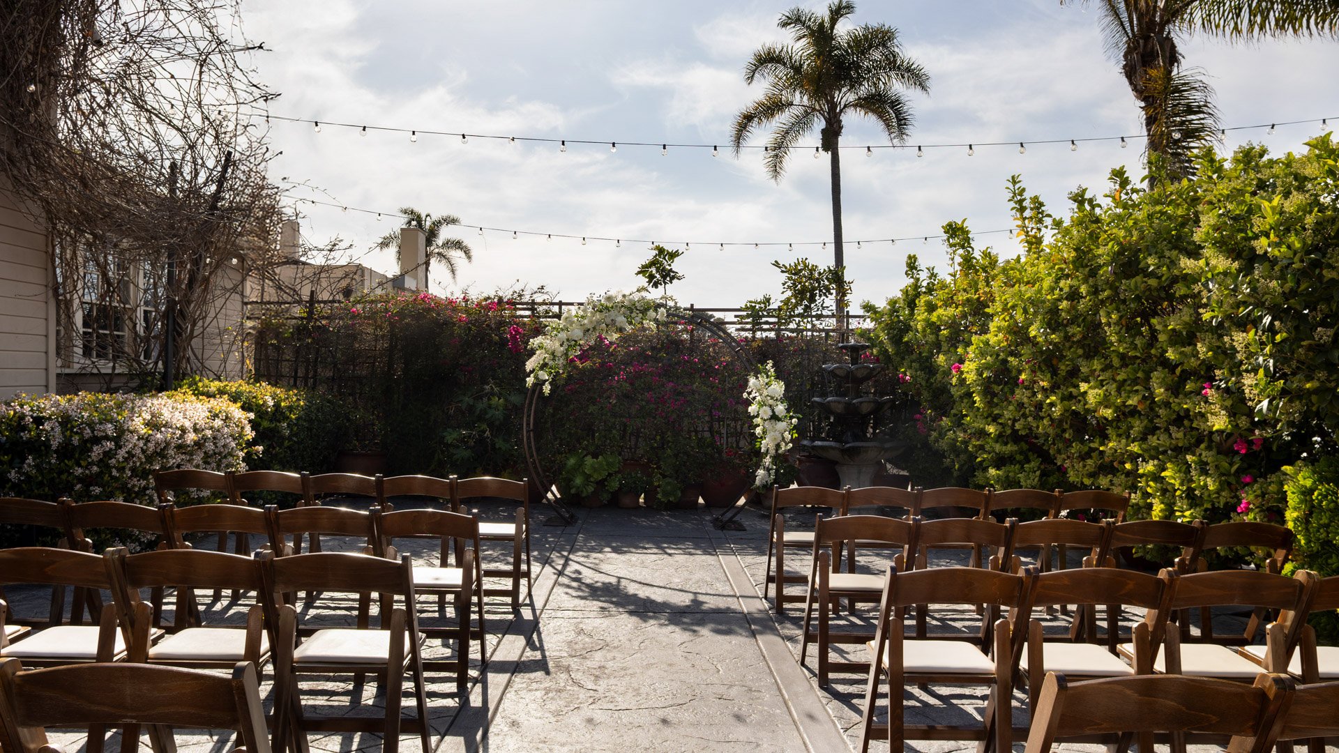Beautiful outdoor wedding ceremony in the Garden Courtyard at Cuvier Club featuring white floral arch, wooden cross-back chairs, string lights, and tropical bougainvillea in La Jolla