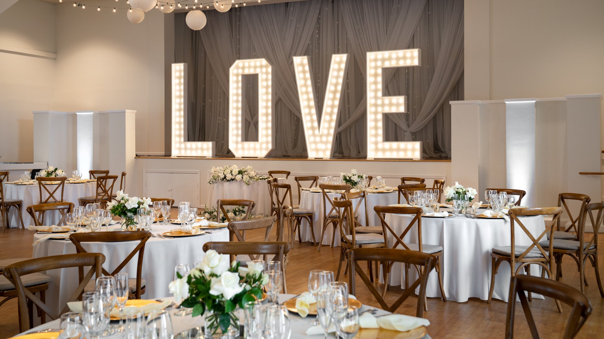 Grand indoor wedding reception space at Cuvier Club by Wedgewood Weddings with illuminated LOVE letters, white uplighting, cross-back chairs, and paper lantern ceiling in La Jolla
