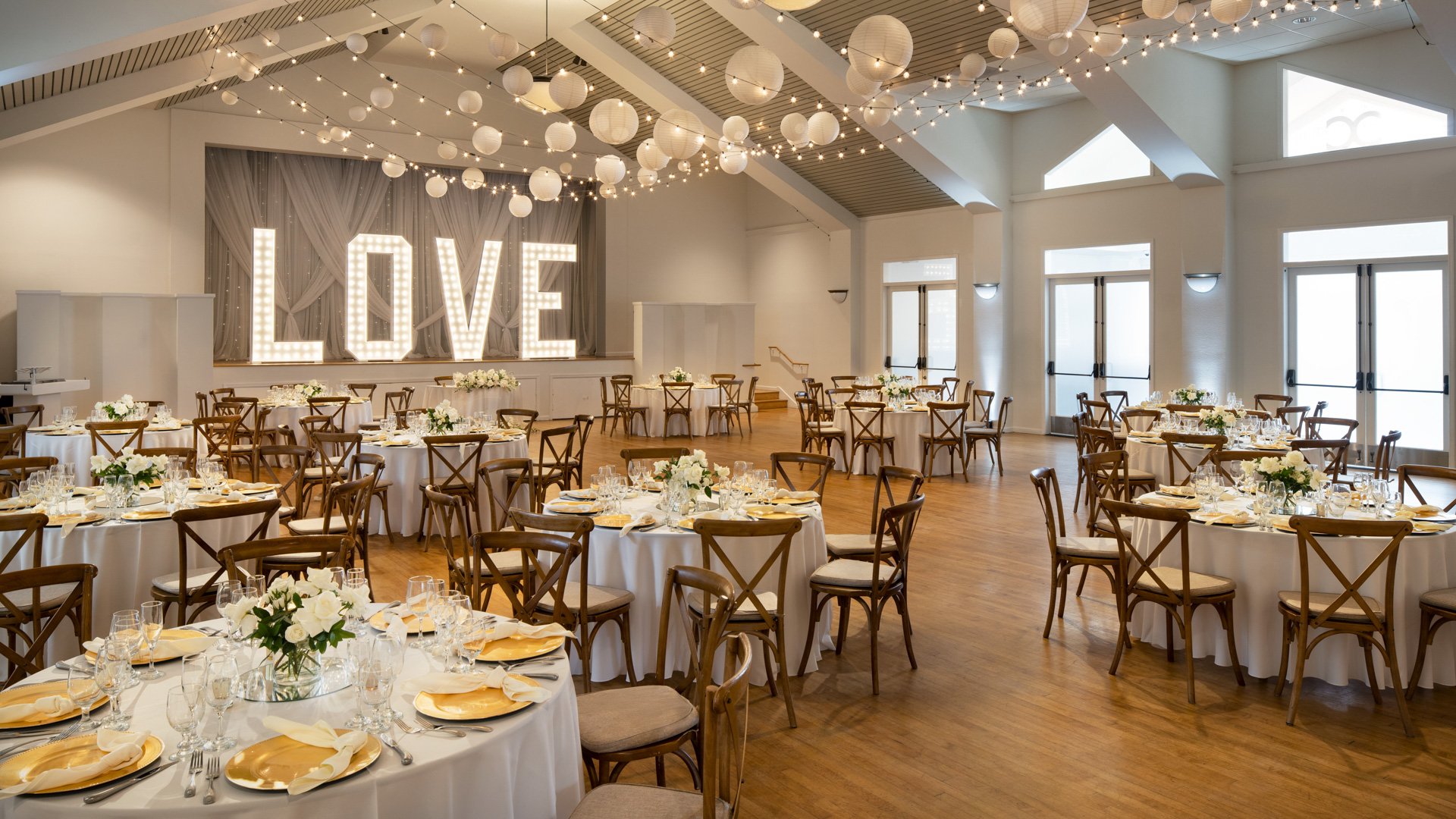 Sophisticated indoor wedding reception space at Cuvier Club La Jolla with cross-back wooden chairs, round tables with white linens, illuminated LOVE marquee sign, and paper lantern ceiling decor