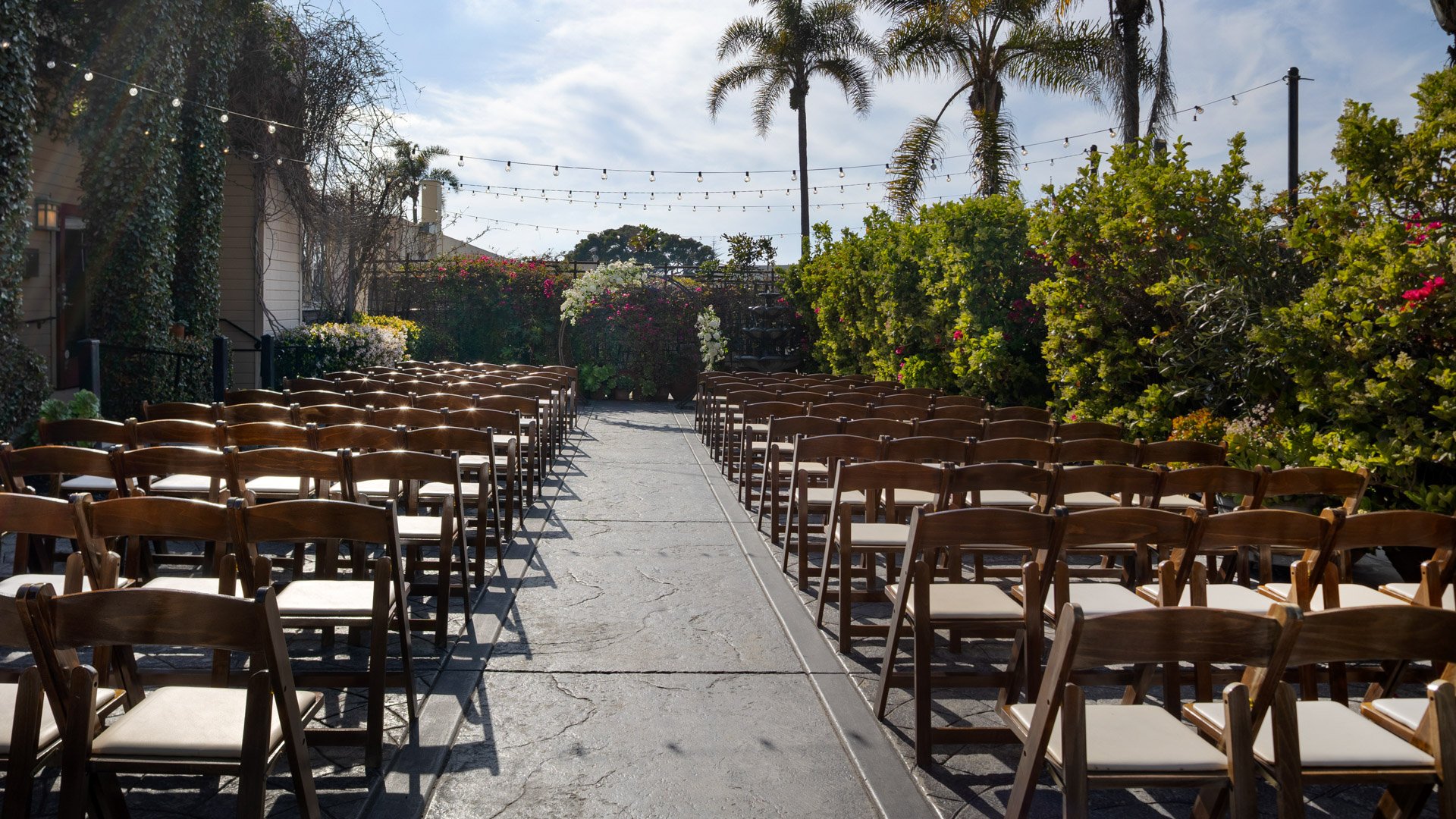 Garden Courtyard ceremony aisle at Cuvier Club by Wedgewood Weddings with wooden cross-back chairs, tropical landscaping, and string lights in La Jolla, California