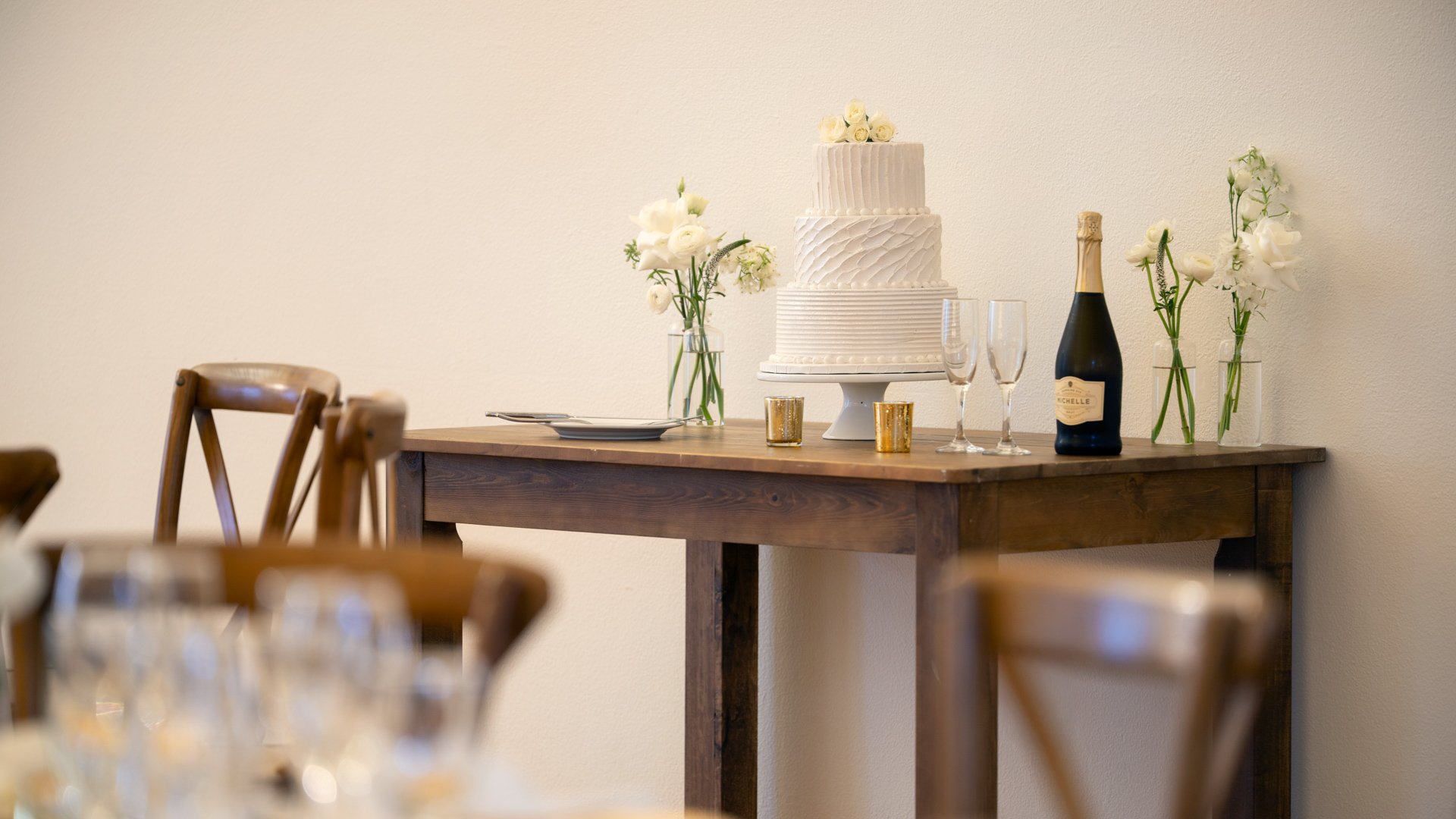Beautiful three-tier white wedding cake display at Cuvier Club featuring textured buttercream design, champagne flutes, white floral arrangements, and rustic wooden table