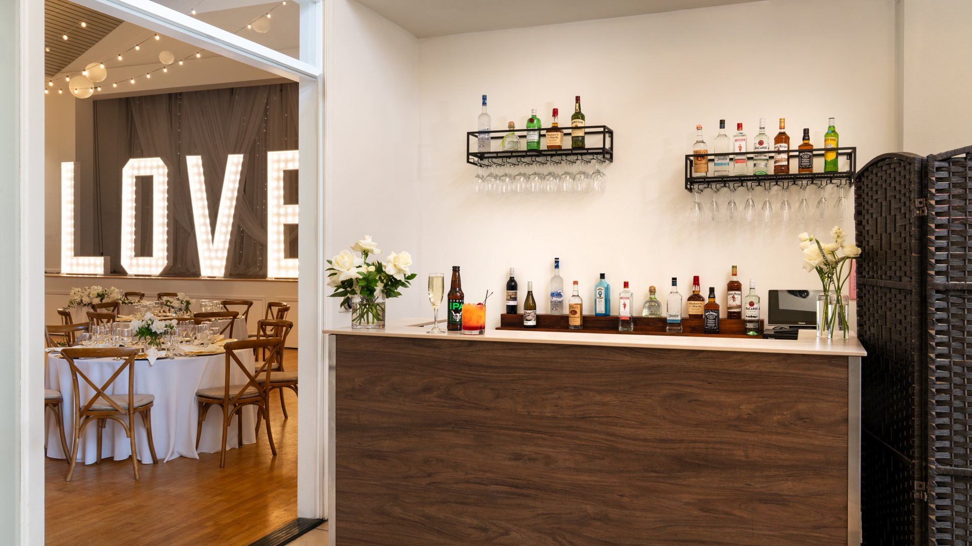 Full-service wedding reception bar at Cuvier Club by Wedgewood Weddings with walnut bar, premium spirits, glassware display, and view into illuminated LOVE sign reception hall