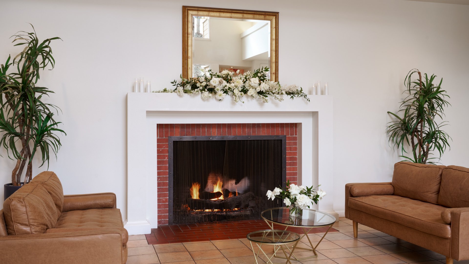 Cozy wedding reception lounge area at Cuvier Club by Wedgewood Weddings featuring brick fireplace, leather armchairs, white floral mantel decor, and tropical plants in La Jolla