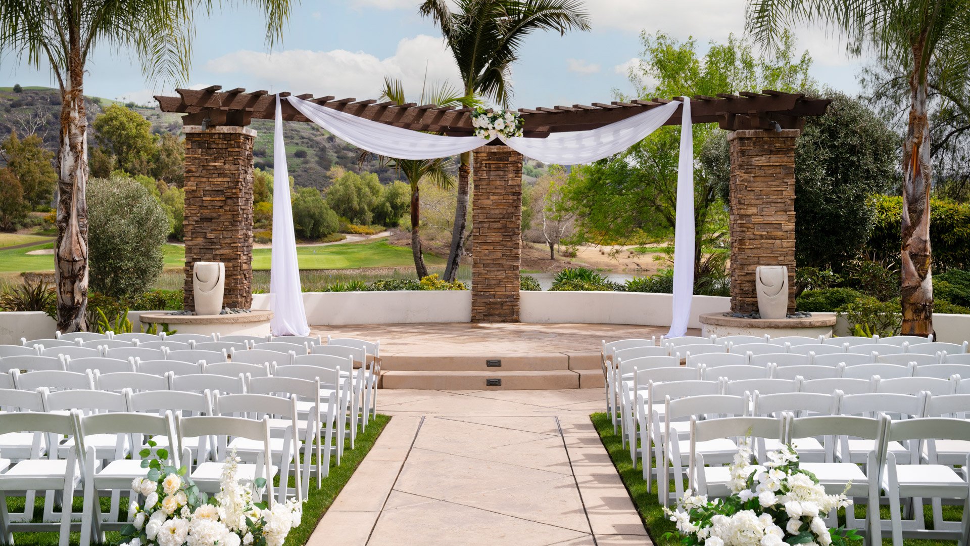 Elegant stone archway wedding ceremony at Fallbrook Estate by Wedgewood Weddings featuring white draping, white folding chairs, floral aisle arrangements and scenic golf course backdrop in Fallbrook California
