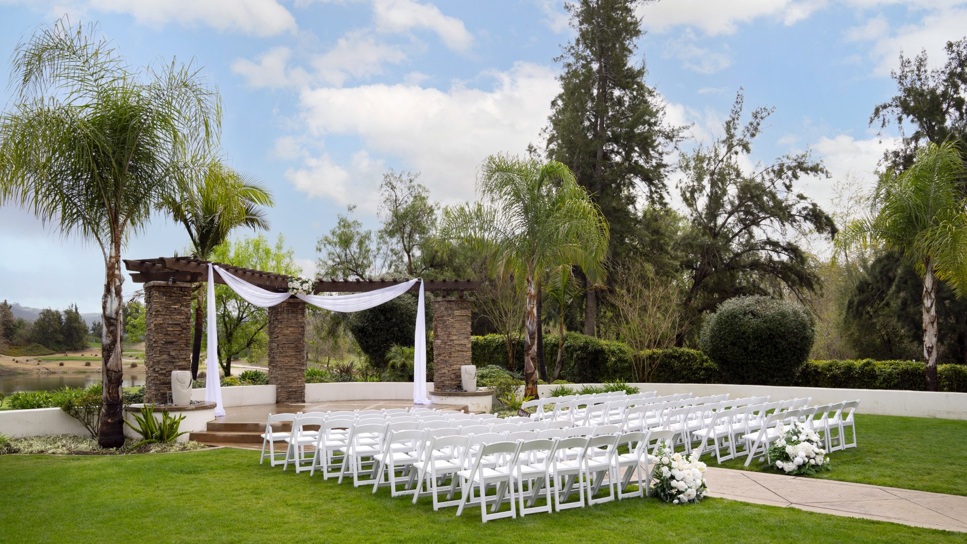 Picturesque garden wedding ceremony at Fallbrook Estate Wedgewood Weddings with stone archway, white draping, white chairs on manicured lawn and tropical palm trees