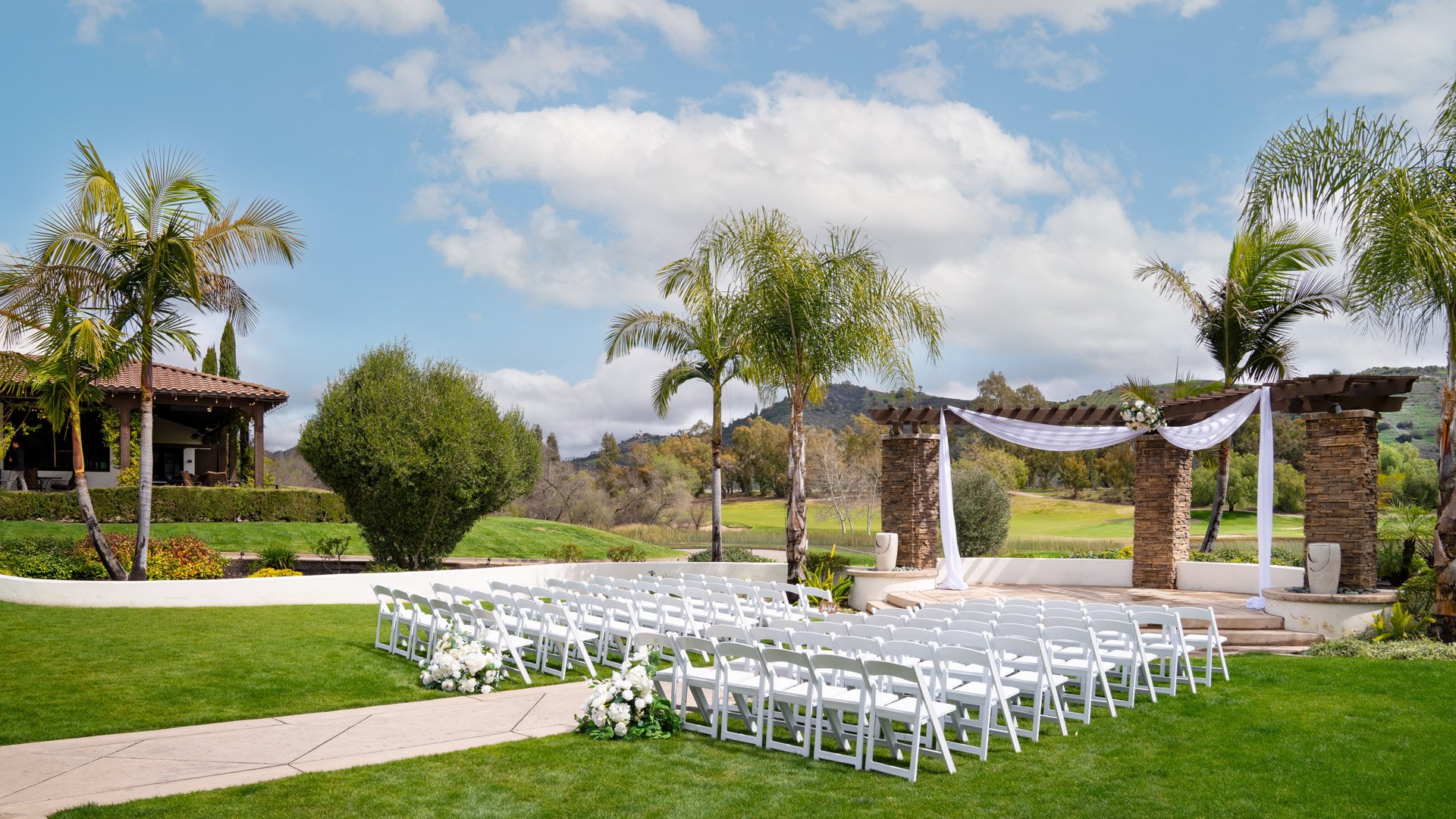 Picturesque garden wedding ceremony at Fallbrook Estate with white chairs on manicured lawn, stone archway backdrop, rolling hills and golf course views in Fallbrook California
