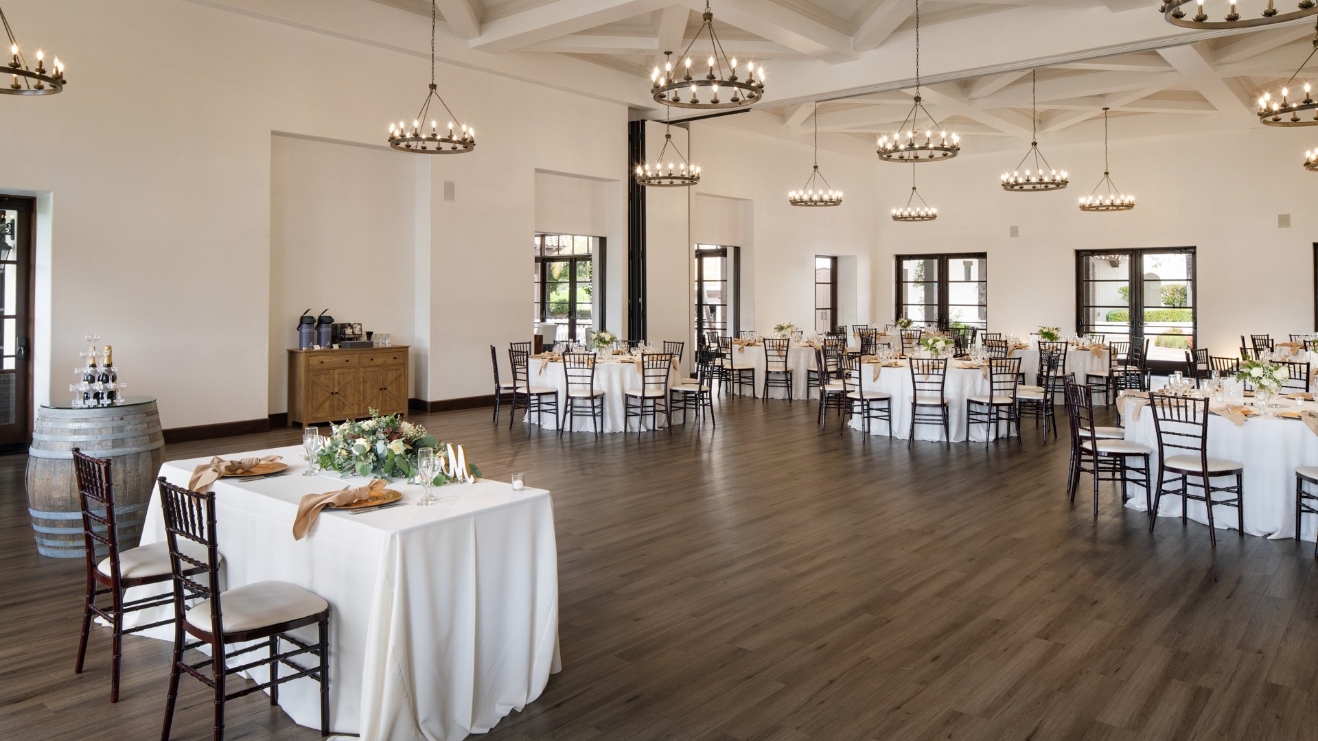 Spacious wedding reception ballroom at Fallbrook Estate Wedgewood Weddings with coffered ceiling, wrought iron chandeliers, round tables with Chiavari chairs, and wine barrel accent