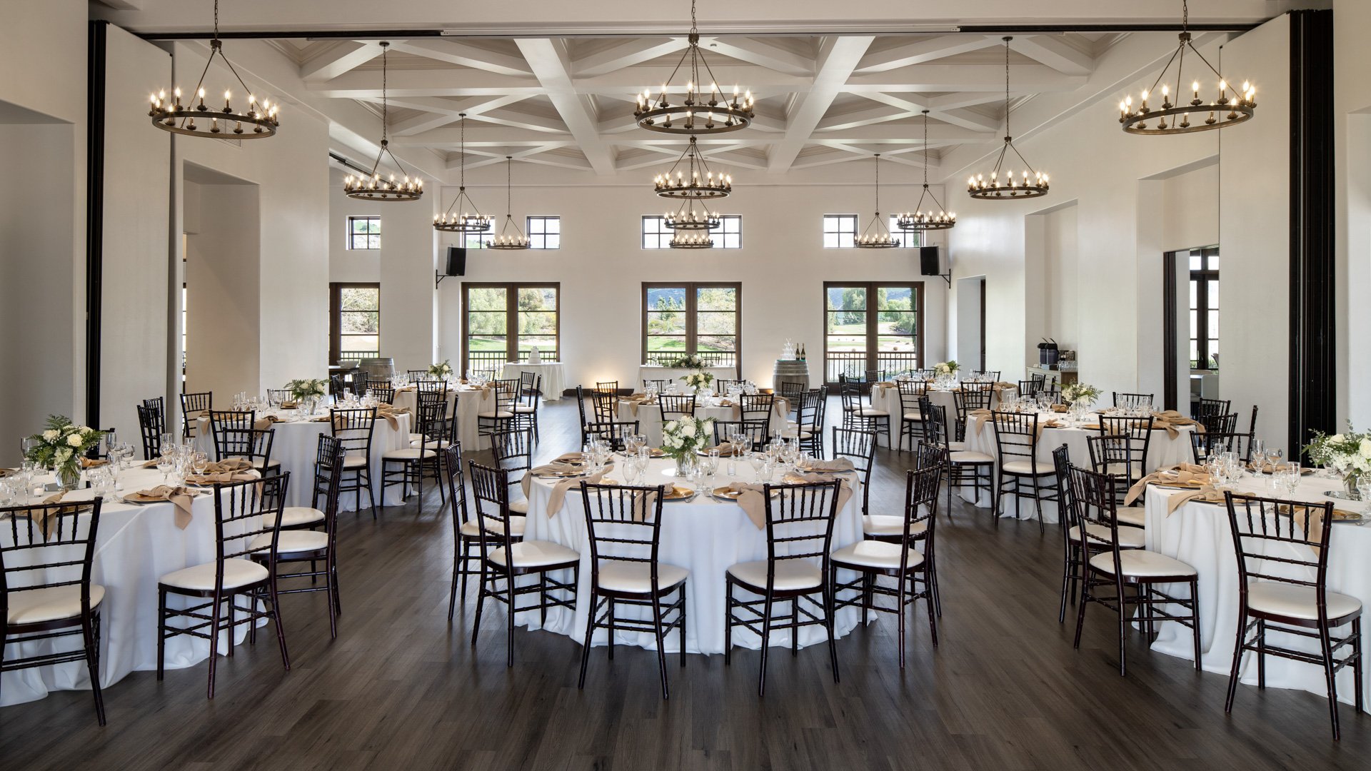 Sophisticated indoor wedding reception space at Fallbrook Estate Wedgewood Weddings featuring exposed beam ceiling, wrought iron chandeliers, round tables with Chiavari chairs and panoramic windows