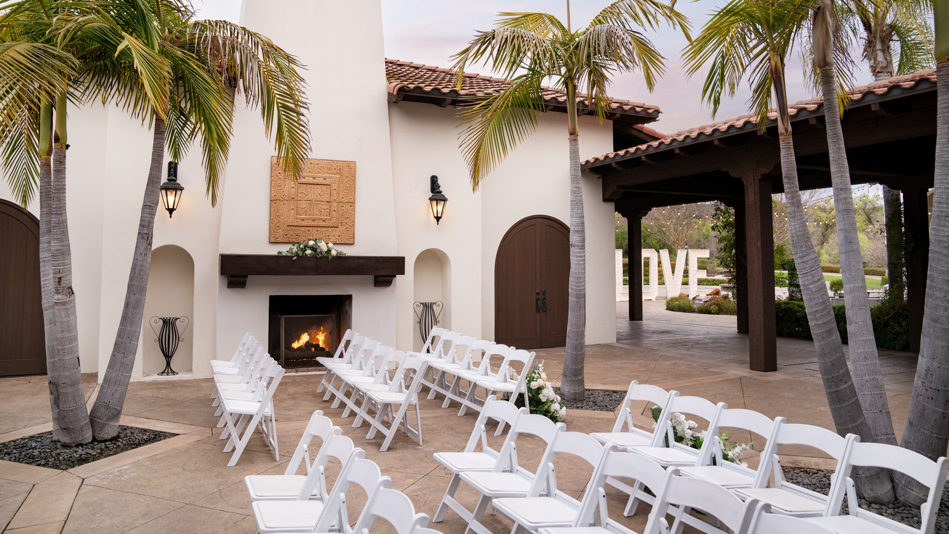 Romantic courtyard wedding ceremony at Fallbrook Estate featuring outdoor fireplace, palm trees, white folding chairs, LOVE sign and Spanish colonial architecture in Fallbrook California
