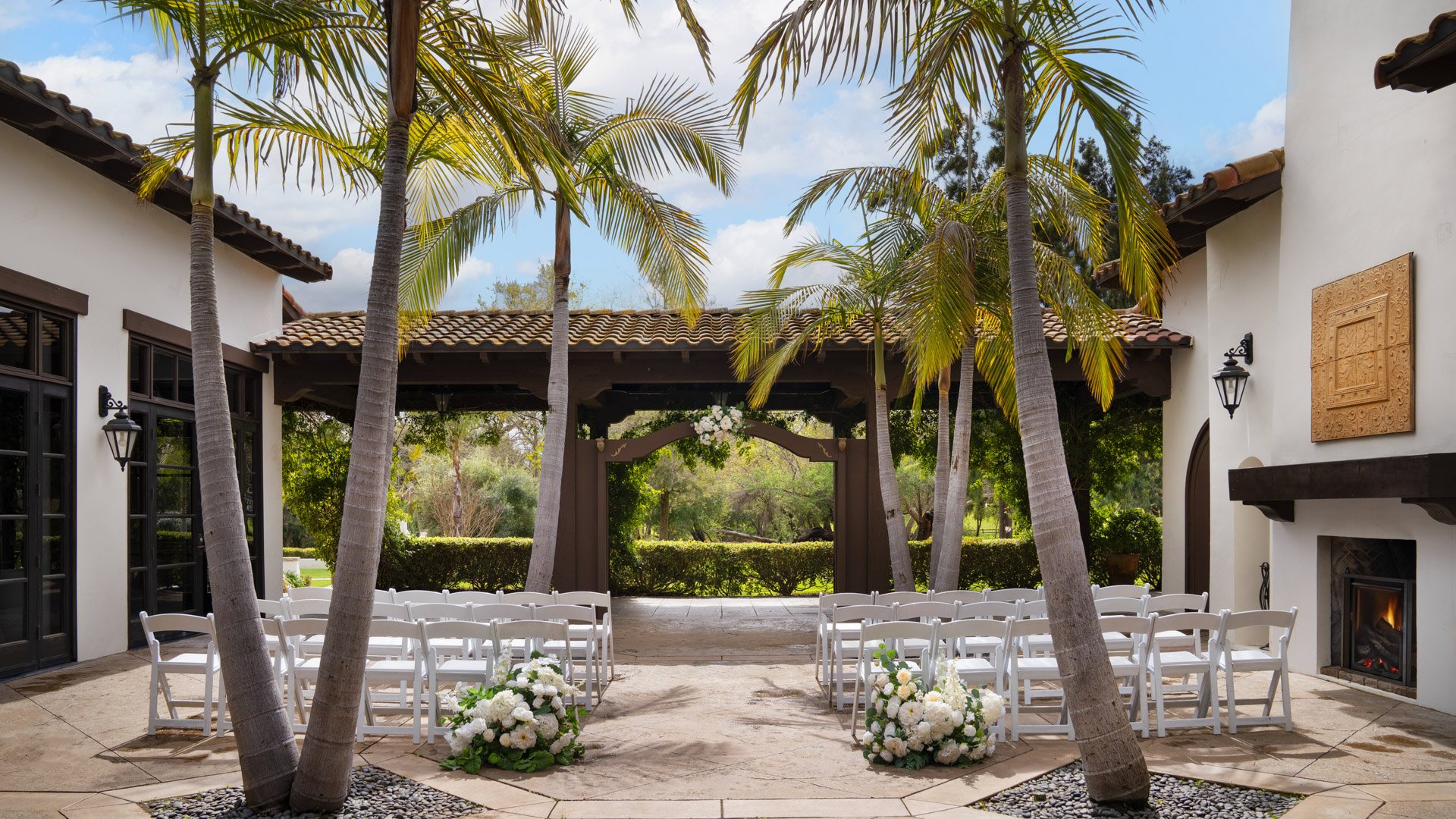 Charming courtyard wedding ceremony at Fallbrook Estate Fallbrook California with white folding chairs, tropical palm trees, Spanish colonial architecture and outdoor fireplace