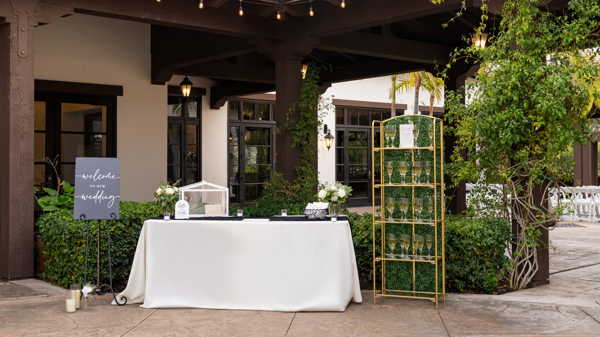 Beautiful outdoor wedding welcome station at Fallbrook Estate featuring welcome signage, gold geometric card display, covered patio with string lights and lush landscaping