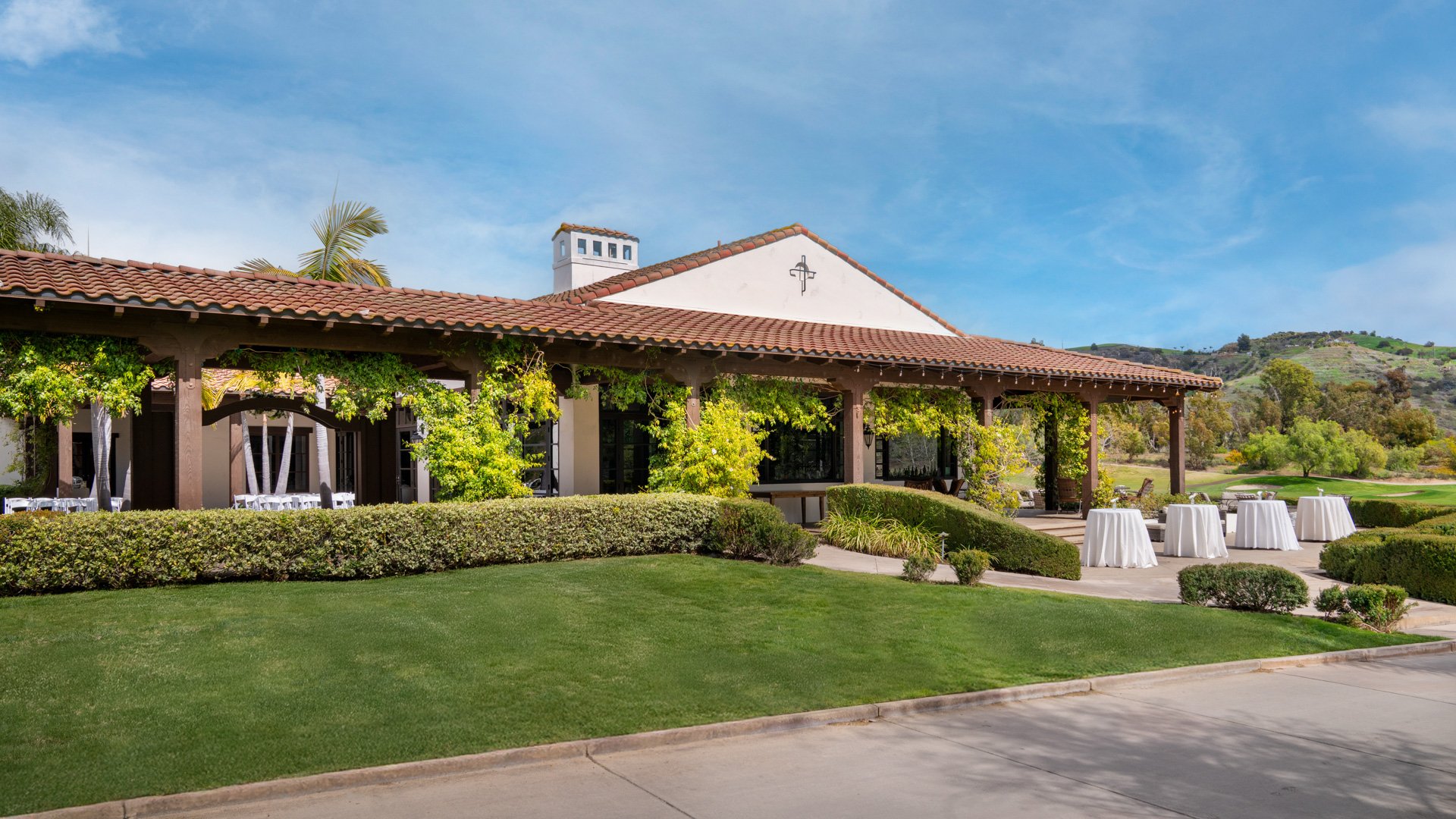 Outdoor wedding cocktail hour setup at Fallbrook Estate featuring Spanish colonial architecture, covered terrace, cocktail tables and manicured gardens with golf course backdrop