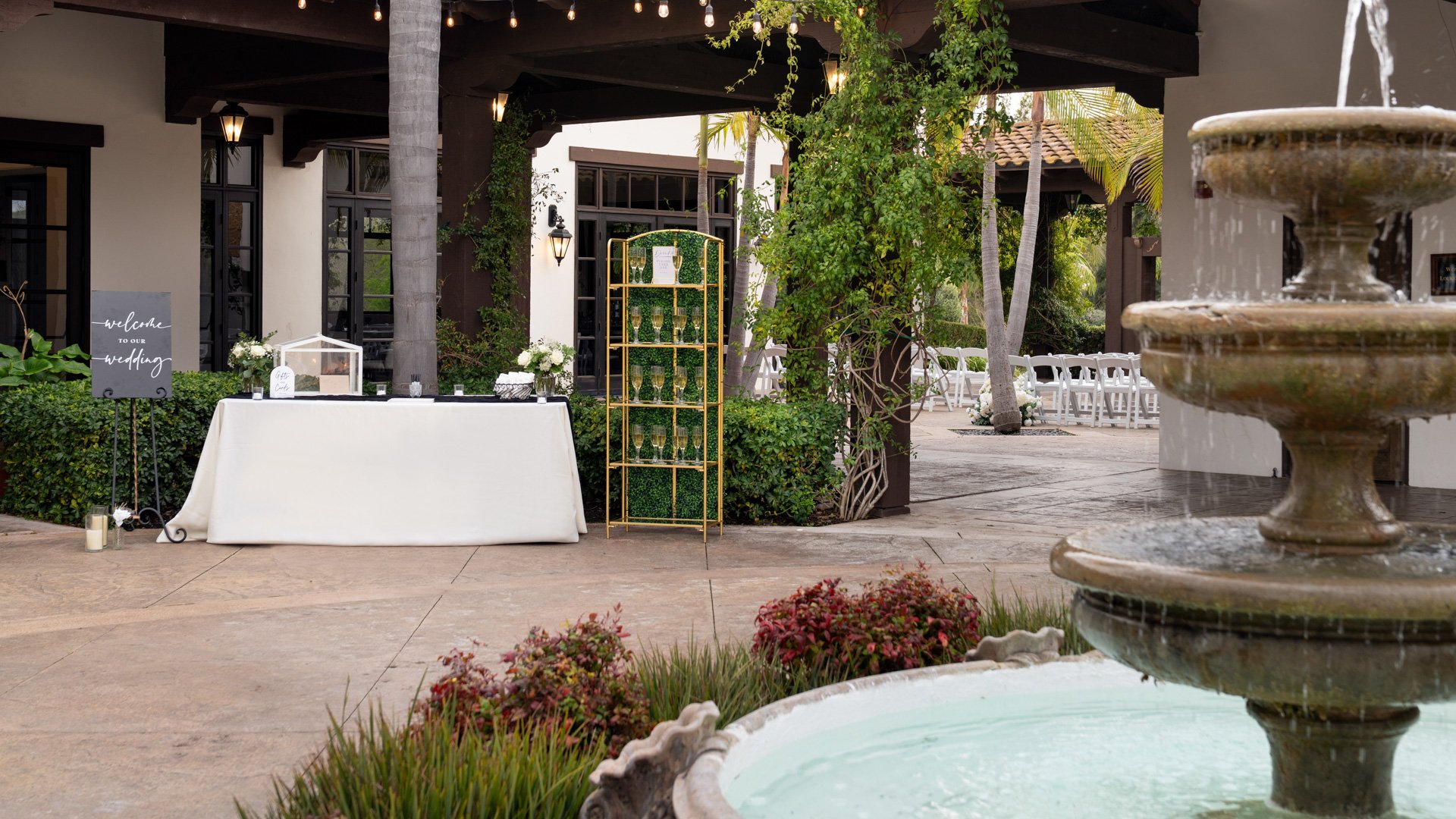 Charming wedding welcome table setup at Fallbrook Estate Wedgewood Weddings with decorative fountain, gold geometric backdrop, covered patio with string lights and Spanish colonial architecture