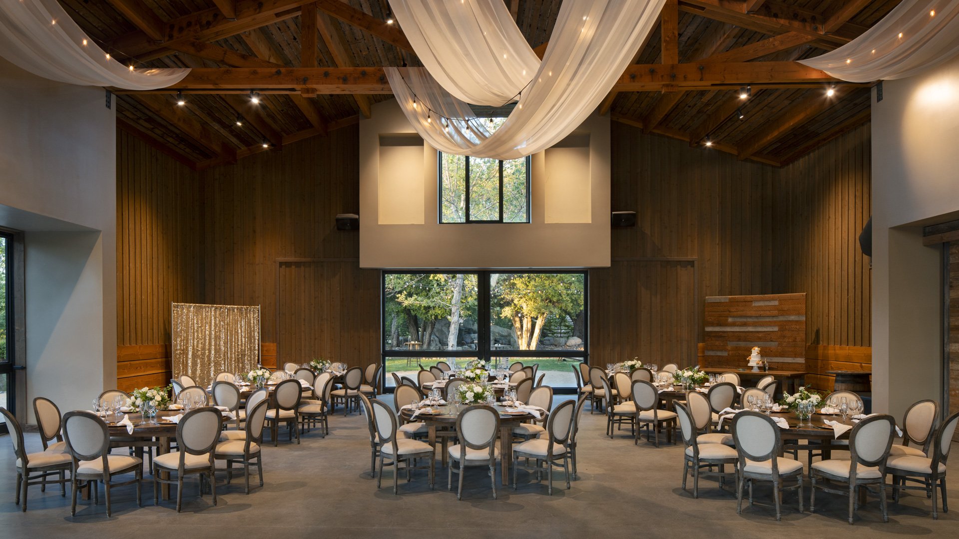 Luxurious reception hall at Galway Downs featuring flowing white fabric ceiling draping with lights, exposed wooden beams, round tables with elegant linens, and garden views through floor-to-ceiling windows in Temecula