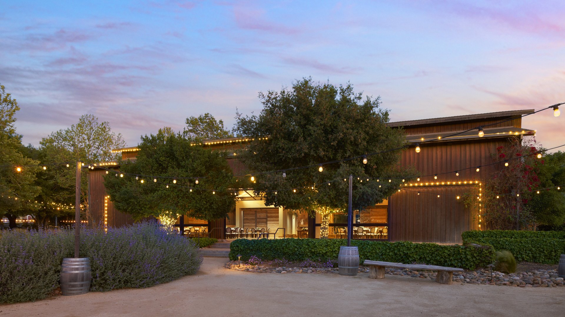Stunning twilight view of Galway Downs rustic wedding venue featuring wooden barn architecture, twinkling string lights, purple sunset skies, and lavender landscaping in Temecula wine country