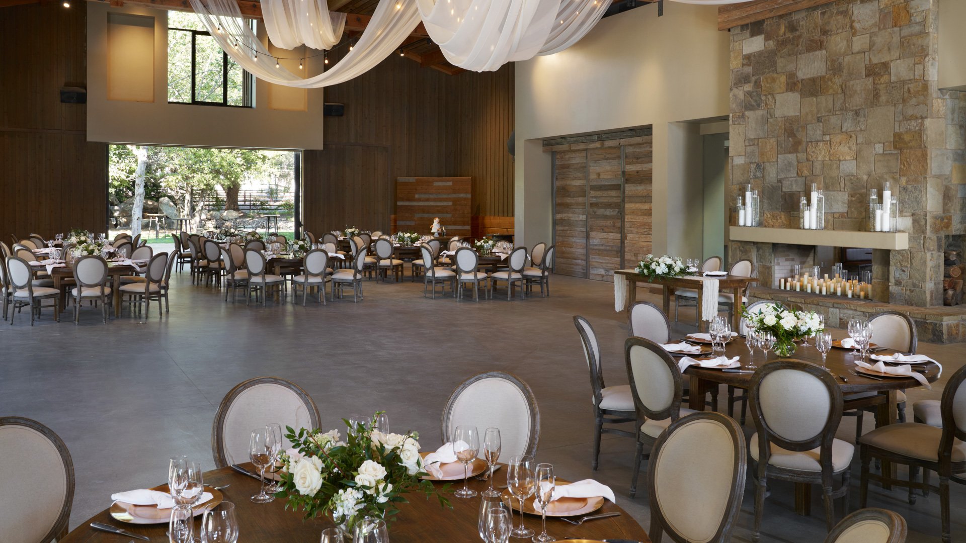 Spectacular grand reception hall at Galway Downs featuring dramatic draped ceiling with twinkling lights, exposed wooden beams, stone fireplace, elegant round tables, and panoramic garden views in Temecula, California