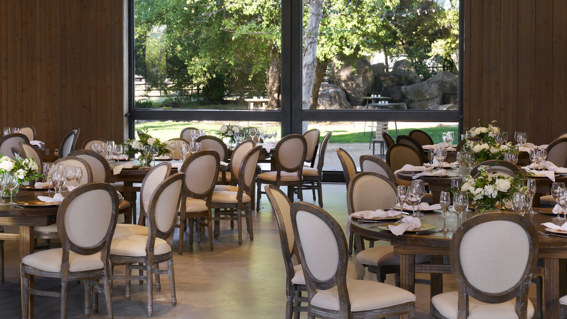 Elegant reception dining at Galway Downs with large windows providing natural light, round tables with white floral centerpieces, neutral chair covers, and garden stone wall views in Temecula, California