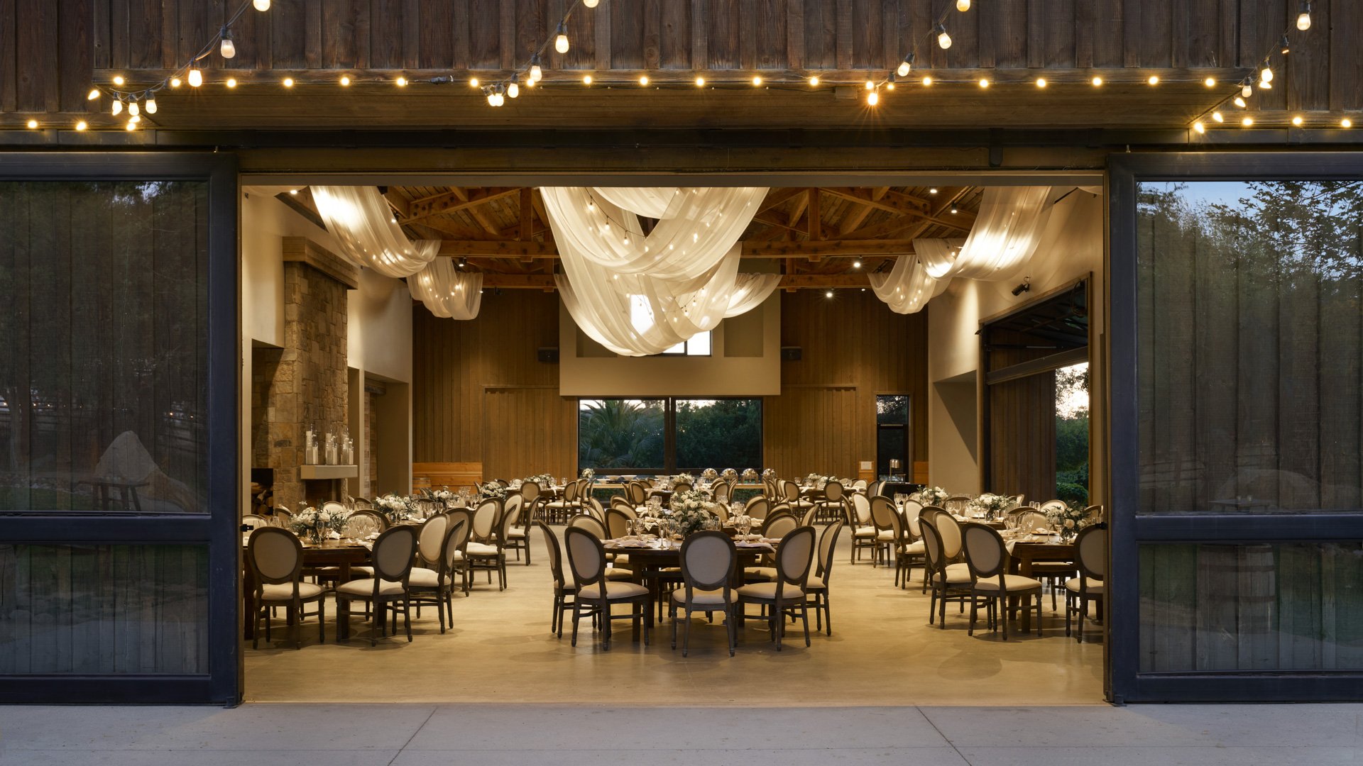 Elegant evening reception at Galway Downs viewed through open barn doors, showcasing draped ceiling lighting, round tables with neutral linens, and warm interior ambiance perfect for Temecula weddings