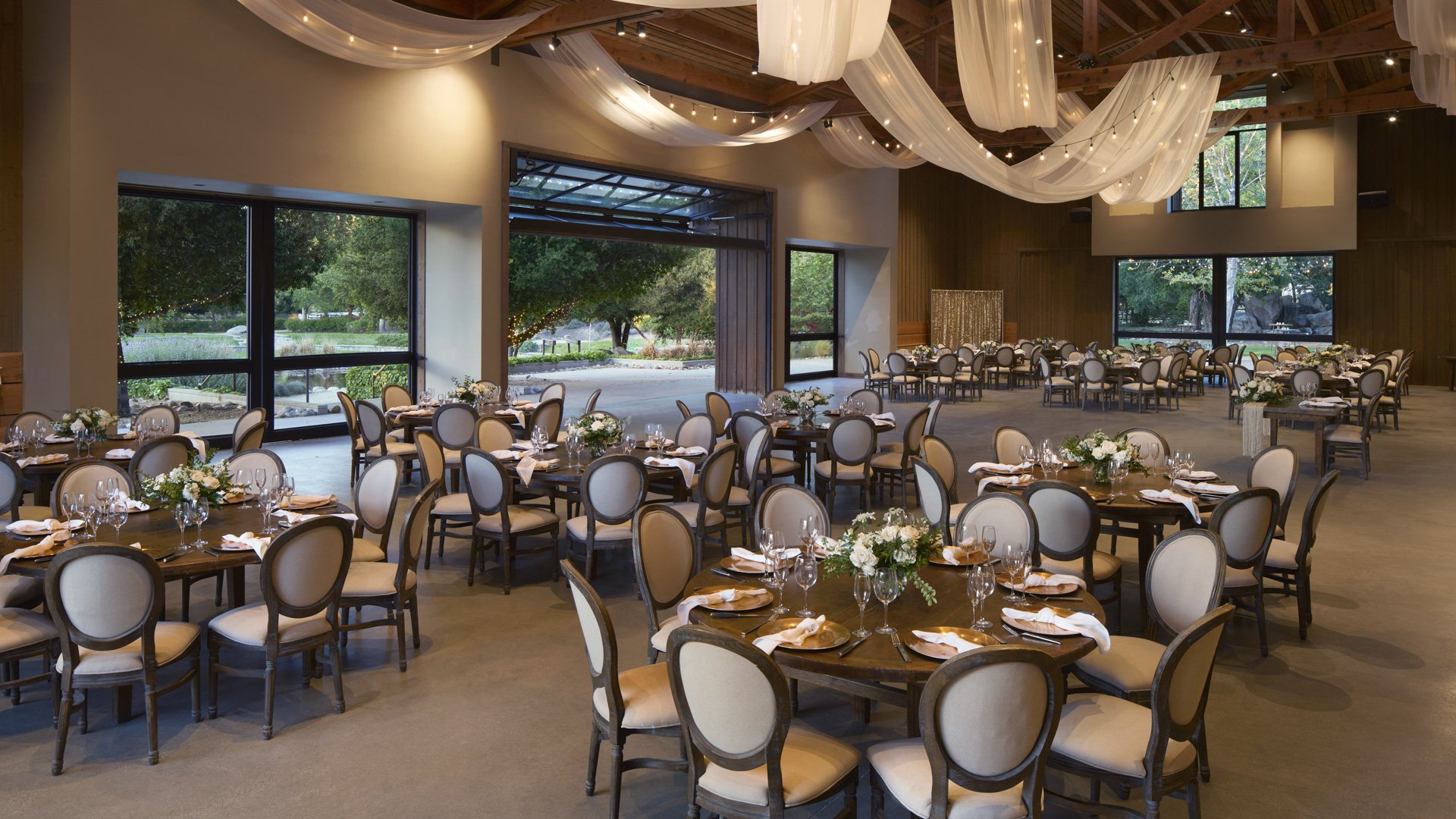Spectacular reception hall at Galway Downs featuring dramatic ceiling draping with twinkling lights, elegant round tables, neutral linens, and floor-to-ceiling windows showcasing garden views in Temecula