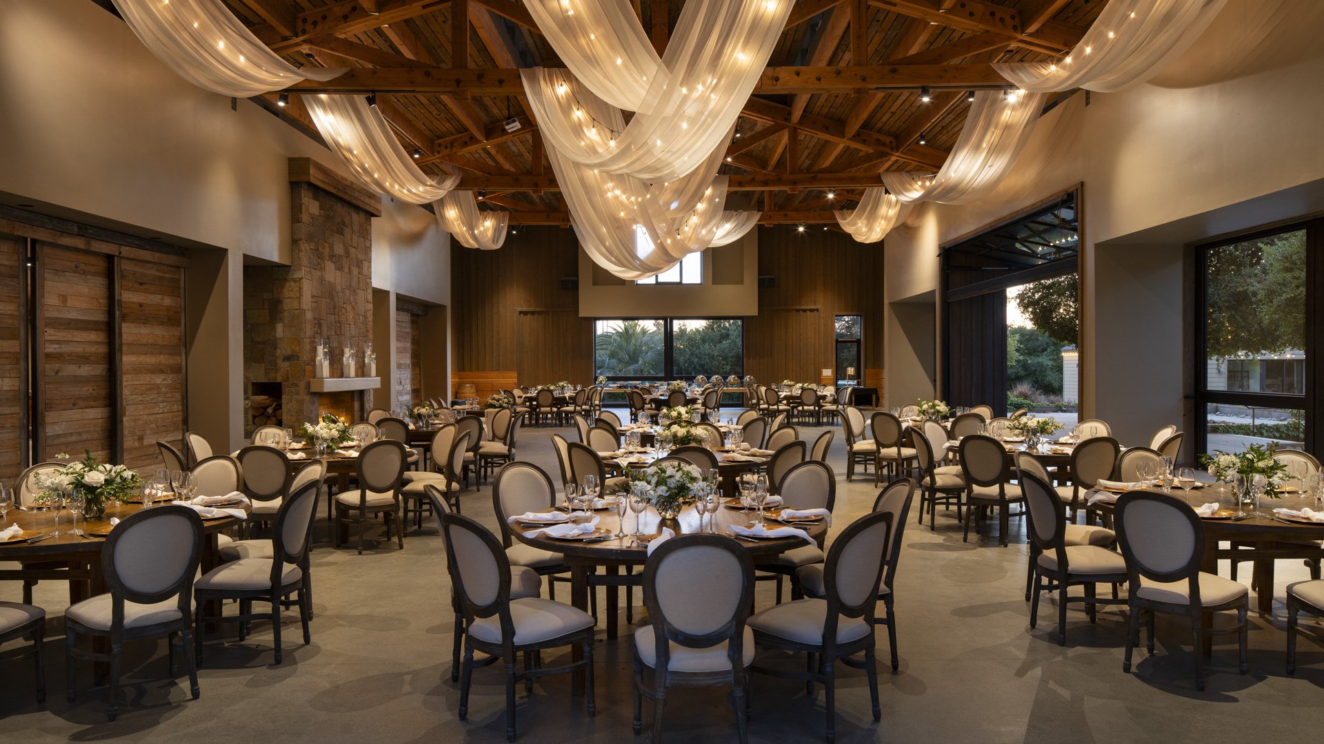 Luxurious indoor wedding reception at Galway Downs featuring exposed wooden beams, flowing white fabric ceiling draping, elegant round tables with neutral linens, and floor-to-ceiling windows overlooking Temecula vineyards