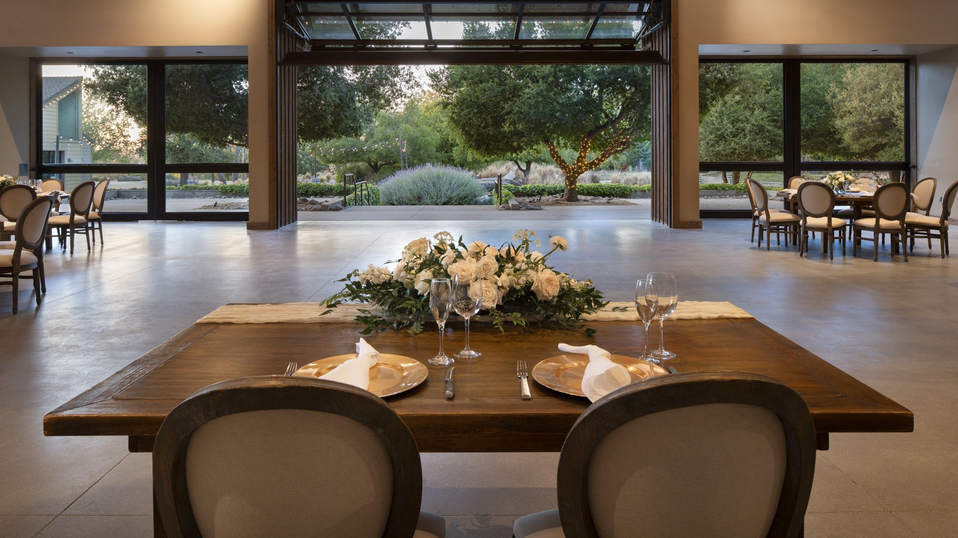 Romantic sweetheart table at Galway Downs featuring rustic wood table, white rose centerpiece, crystal stemware, and garden backdrop through glass walls perfect for intimate wedding dining in Temecula