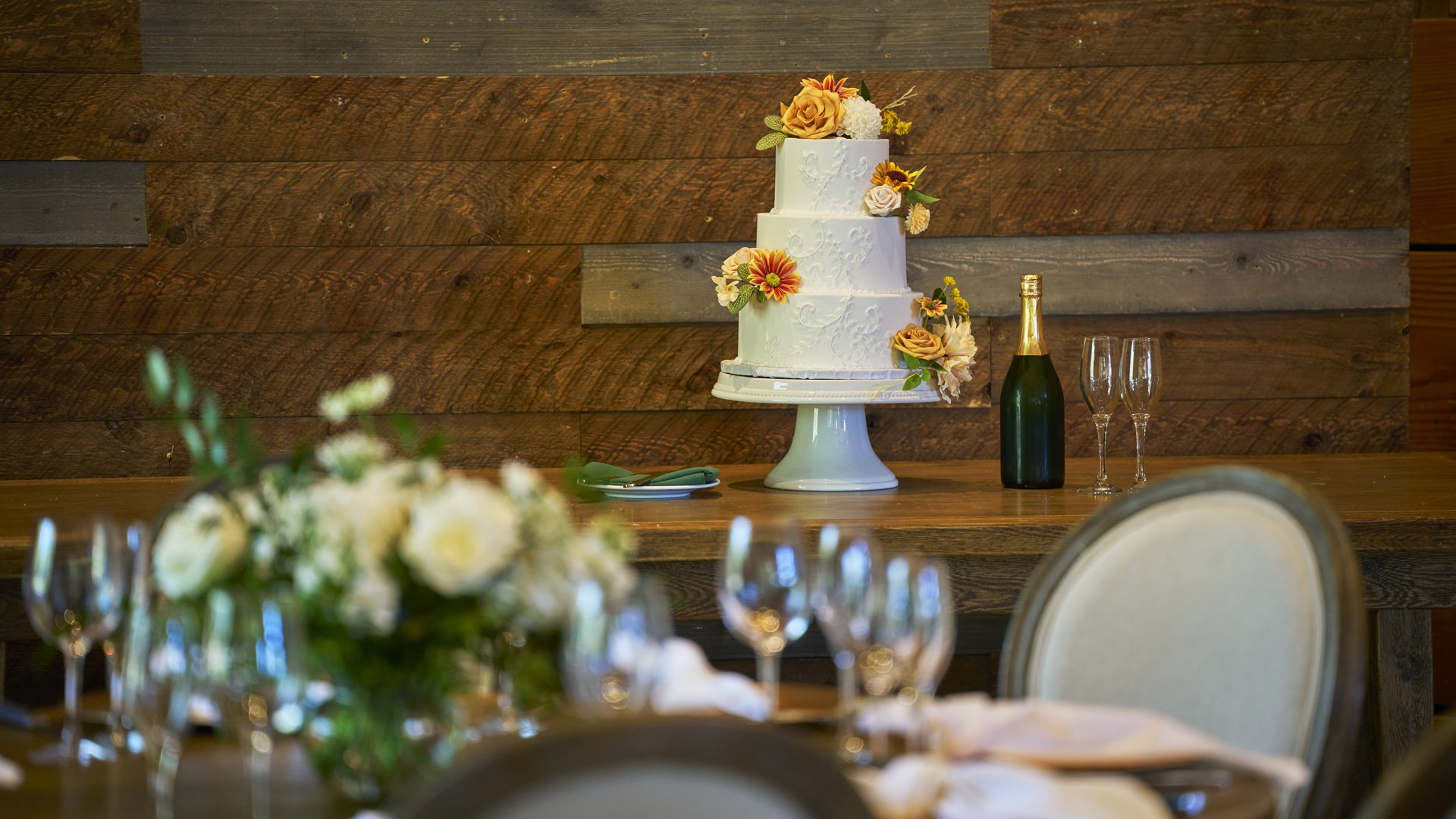 Elegant three-tier wedding cake with autumn floral decorations displayed on rustic wood backdrop at Galway Downs, featuring champagne service and intimate reception setting in Temecula