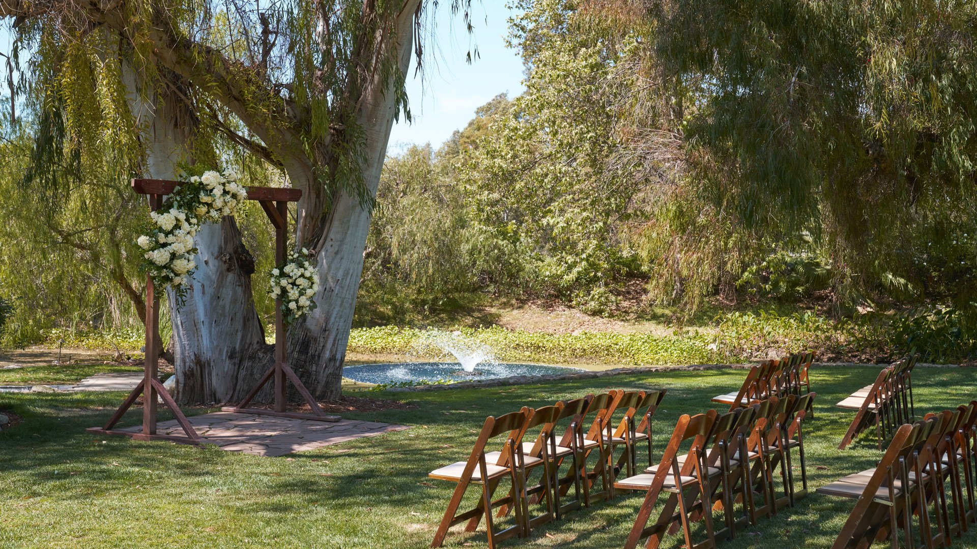 Beautiful lakeside wedding ceremony at The Meadows, Galway Downs featuring rustic wooden arch with white floral arrangements, wooden folding chairs, and serene water feature surrounded by mature trees in Temecula, California
