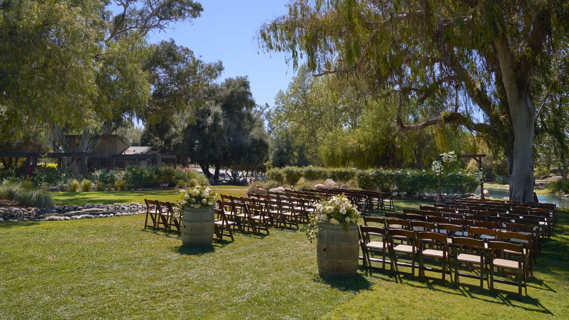 Stunning waterside wedding ceremony at The Meadows, Galway Downs with rustic wooden chairs arranged beside a natural water feature and surrounded by mature trees in Temecula