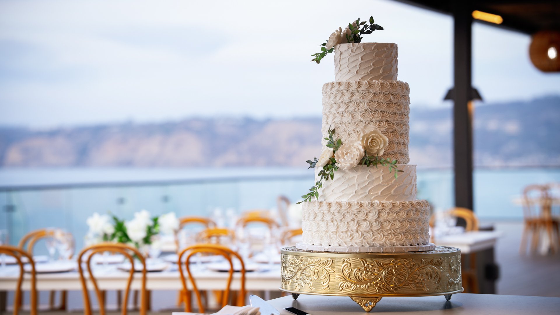 Four-tier white wedding cake with fresh flowers on ornate gold stand, featuring Pacific Ocean views at La Jolla Cove Rooftop wedding venue