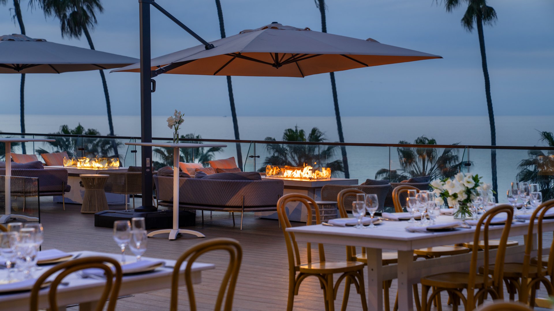 Elegant twilight wedding reception with fire pit lounges, bentwood dining chairs, and glass-enclosed oceanfront terrace at La Jolla wedding venue